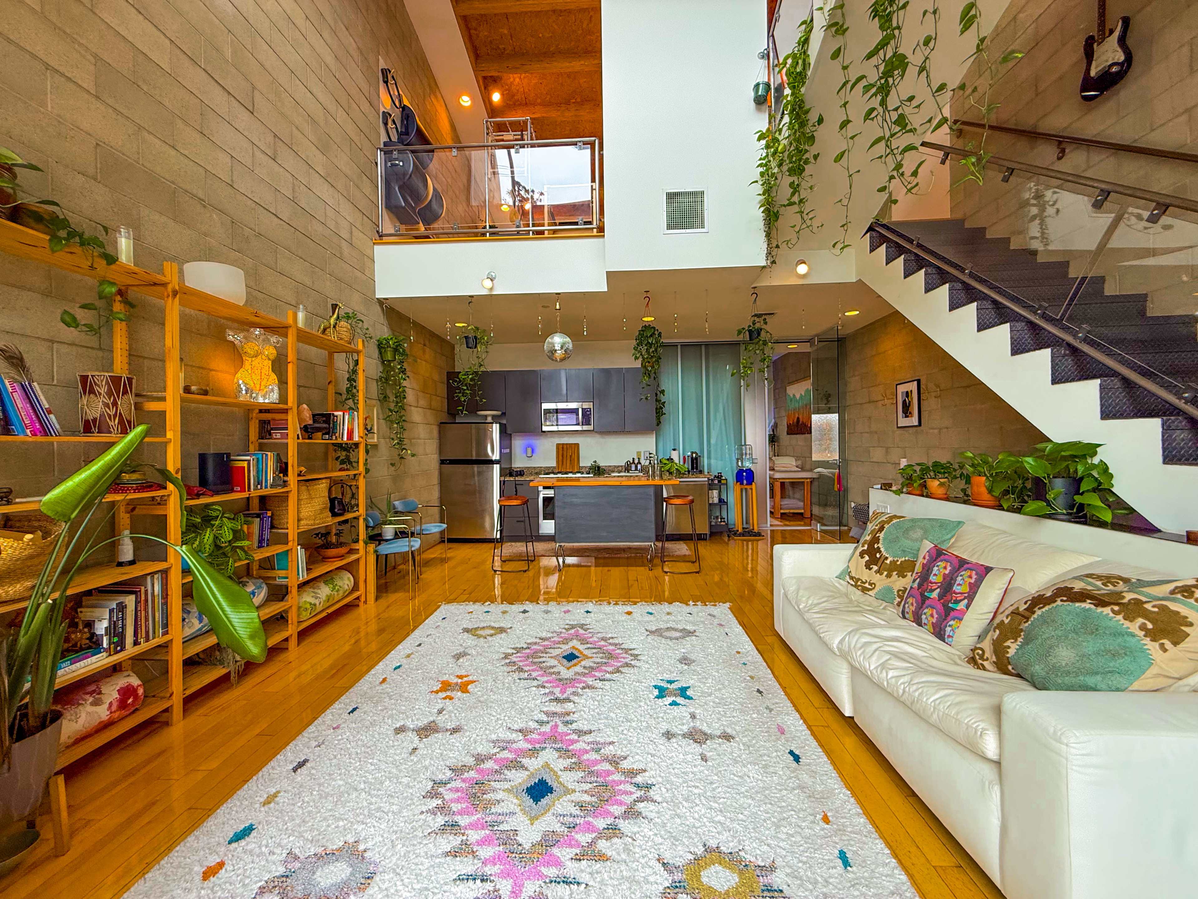 The image shows a modern living room with a white sofa, a large patterned rug, and a staircase leading to an upper level, surrounded by plants and wooden shelving.