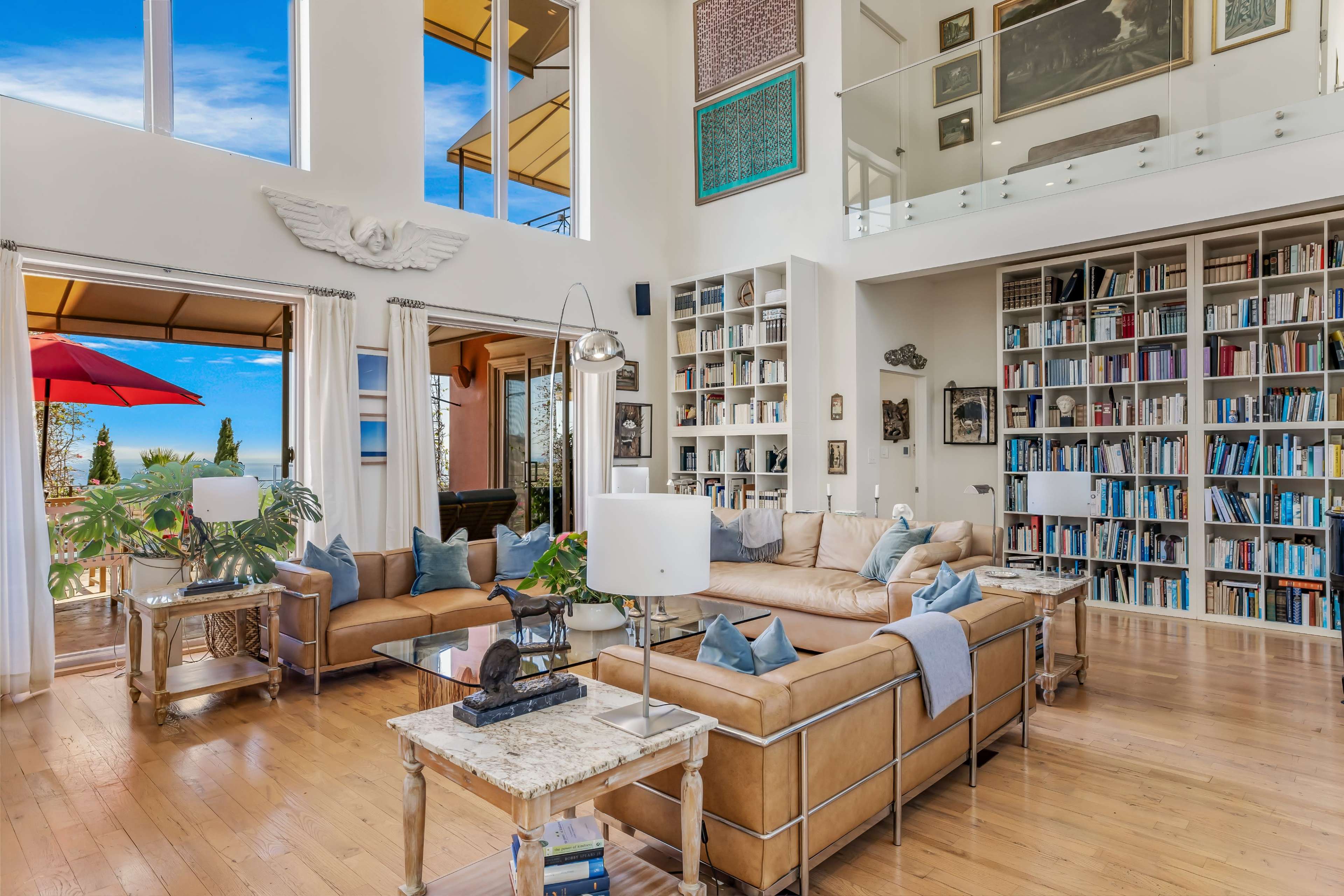 A spacious living room features a large sectional sofa, numerous bookshelves filled with books, and large windows offering a view of the outdoors.