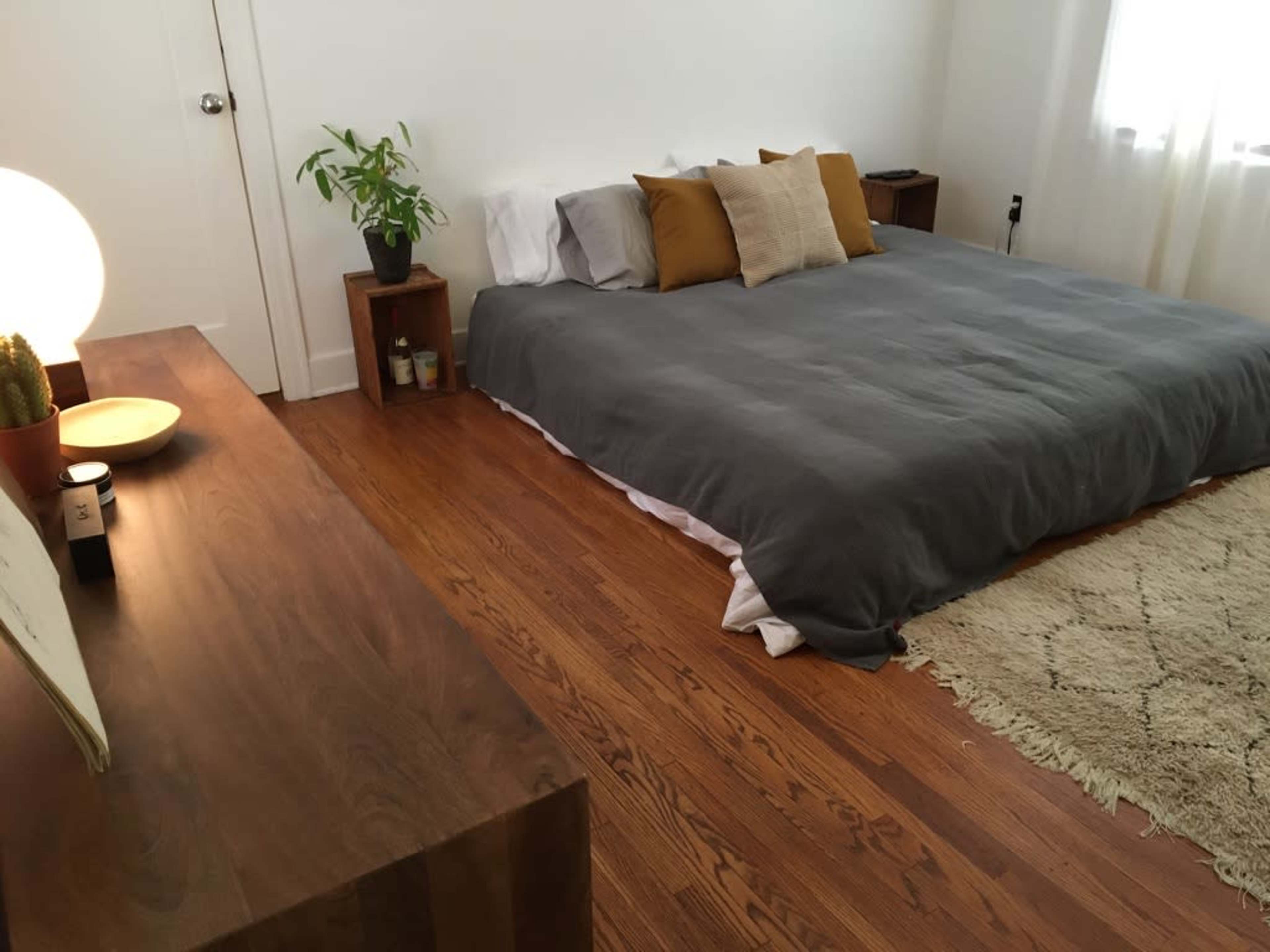 The image shows a minimalist bedroom with a large bed covered in a gray duvet, 
wooden furniture, a plant, and a soft rug on a wooden floor.