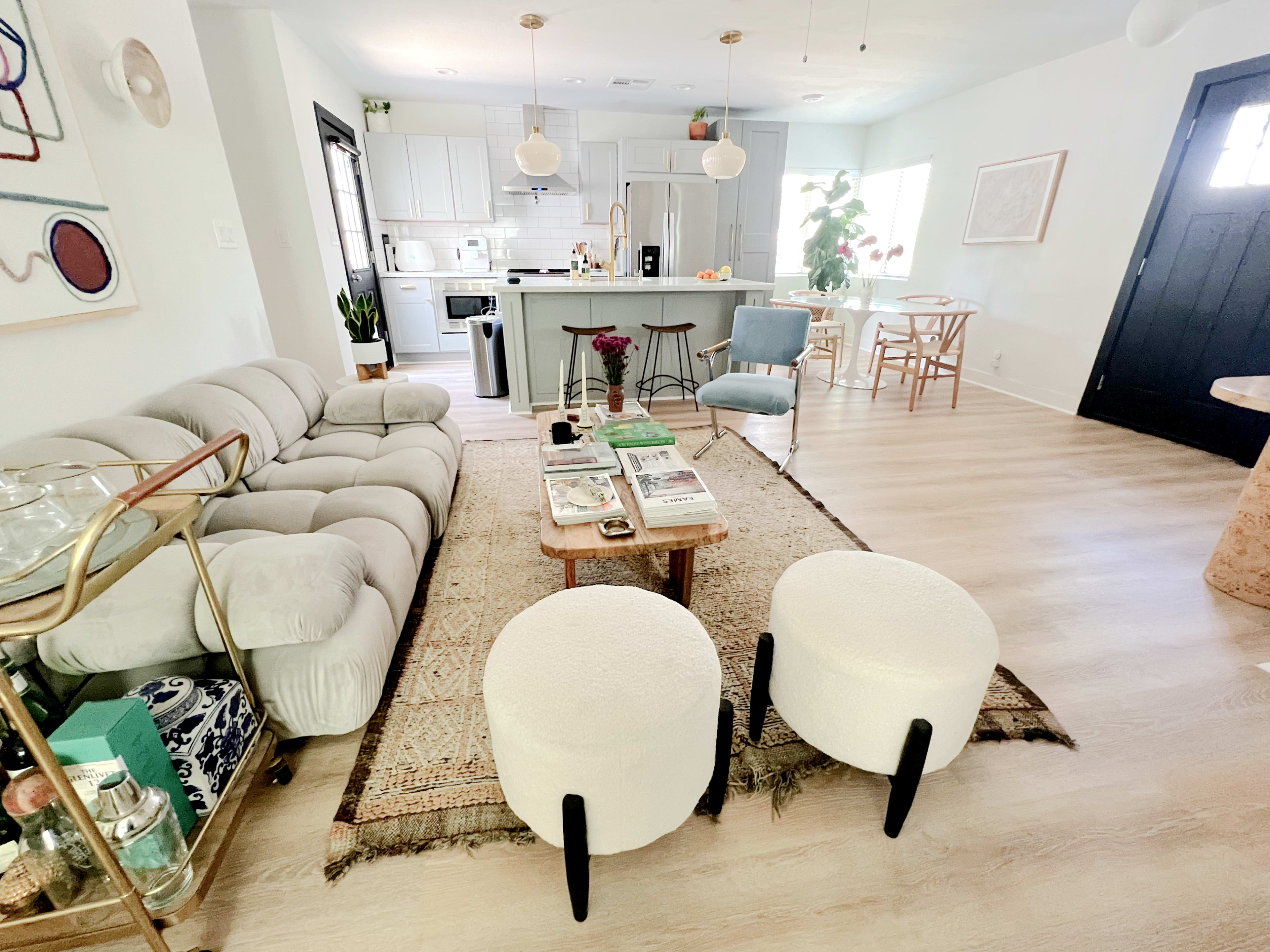 A modern living space with a light-colored sofa, round ottomans, a wooden coffee table with magazines, and a kitchen area in the background.