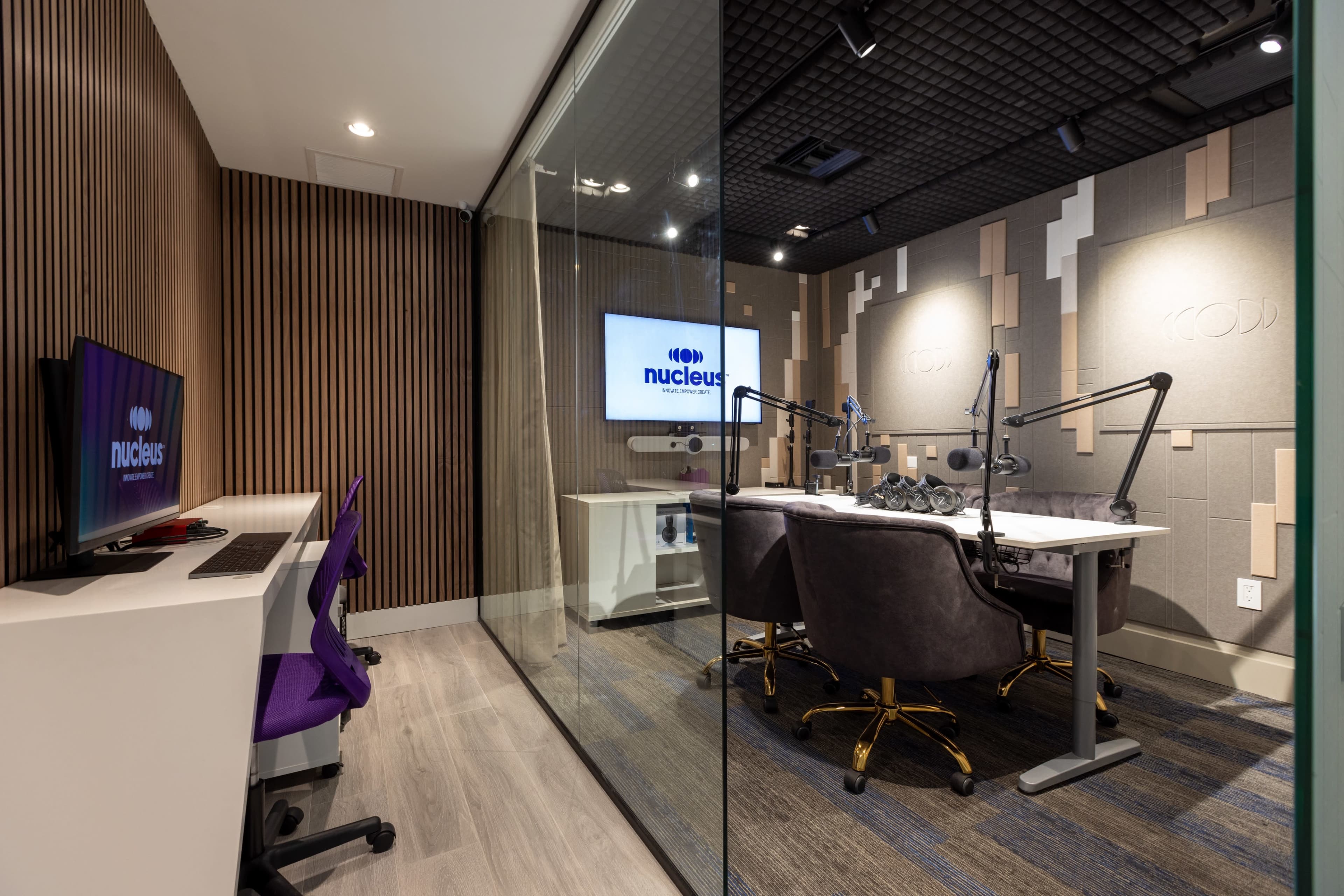 A modern podcast studio features a glass wall, computer desks, and soundproof walls with microphones arranged for recording.