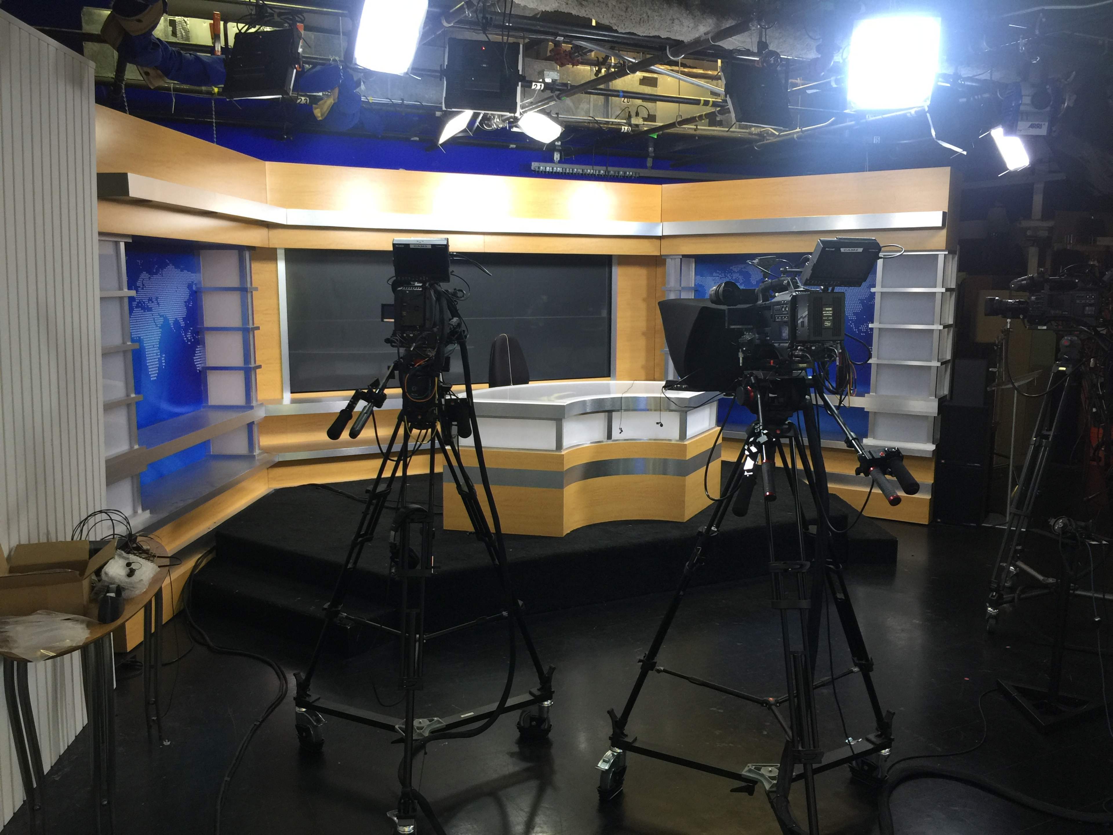 A television news studio is equipped with cameras and lights, featuring a central news desk and a backdrop displaying a world map.