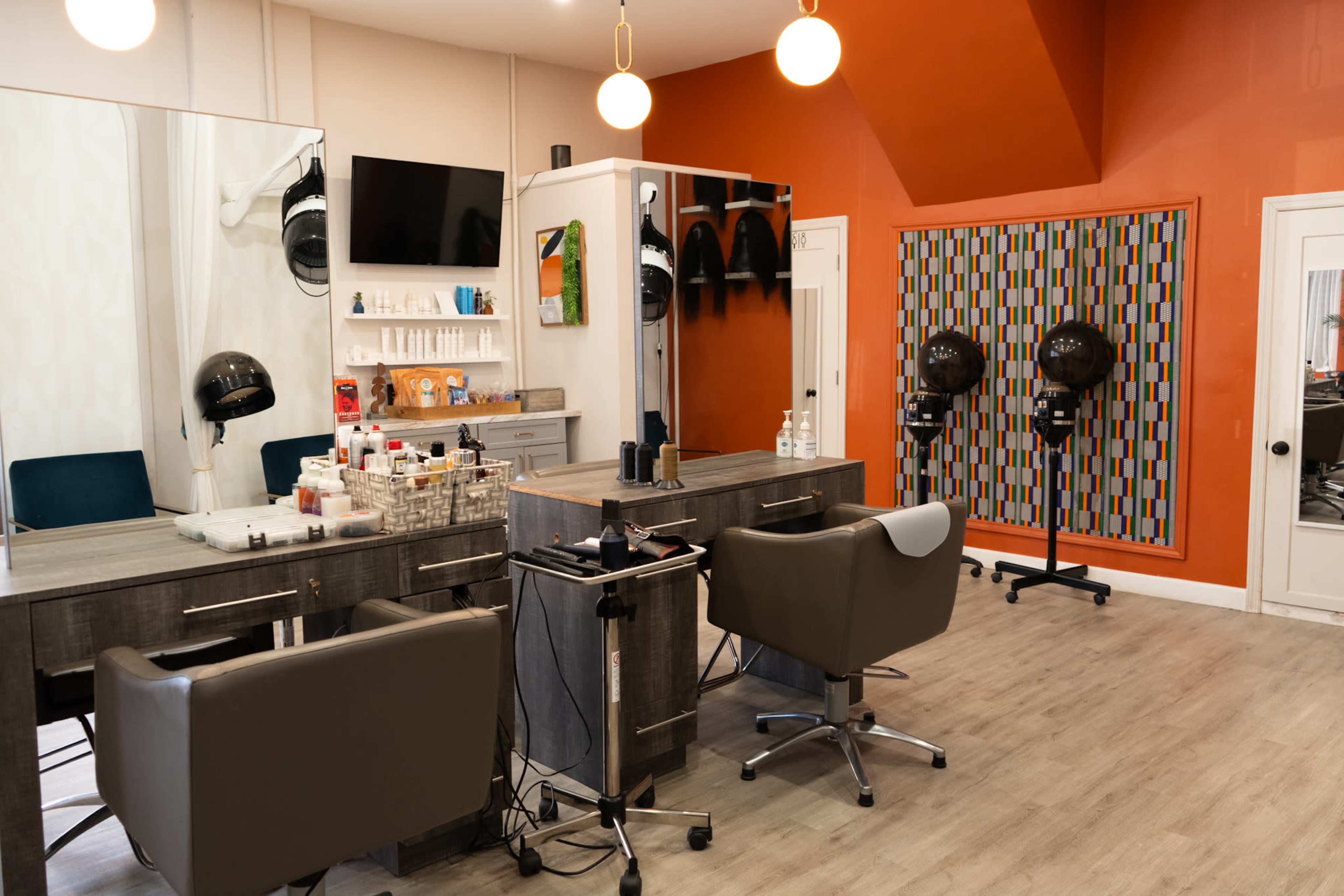 The image shows a modern hair salon featuring two styling stations with chairs, a wall-mounted TV, and hairdryers on stands, all set against an orange accent wall.