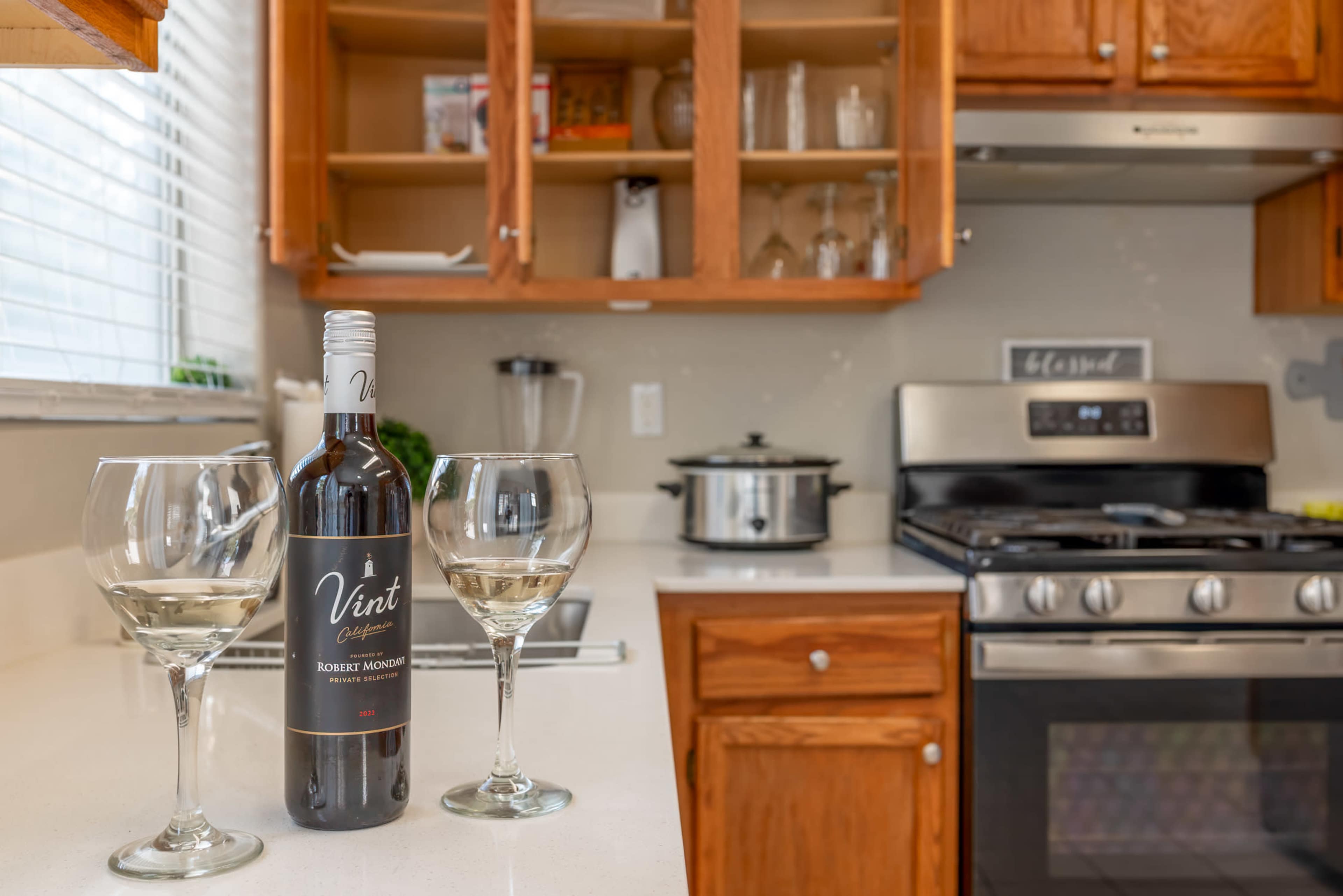 A bottle of wine sits next to two wine glasses on a kitchen countertop with wooden cabinets and a stove in the background.
