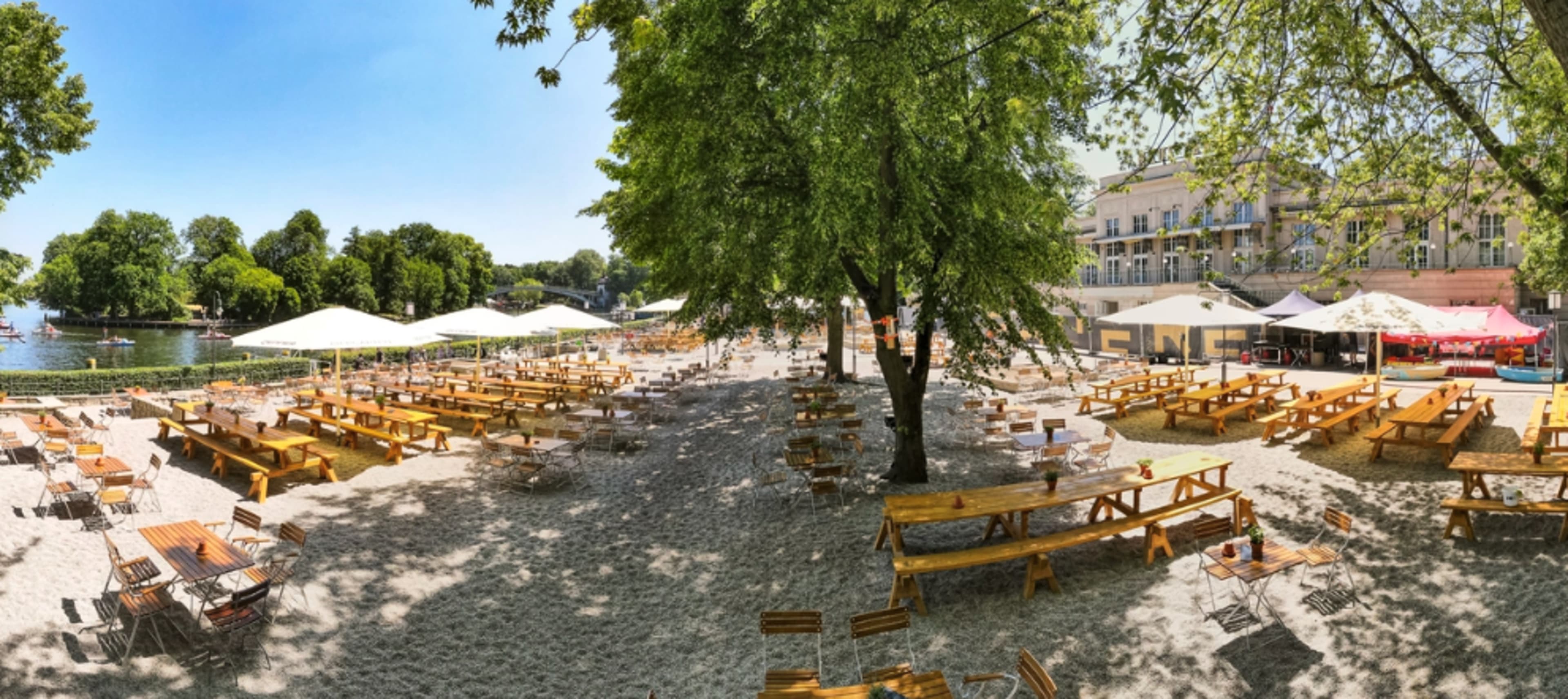 A sandy outdoor area with numerous wooden tables and benches, shaded by large trees, near a body of water and several tents in the background.
