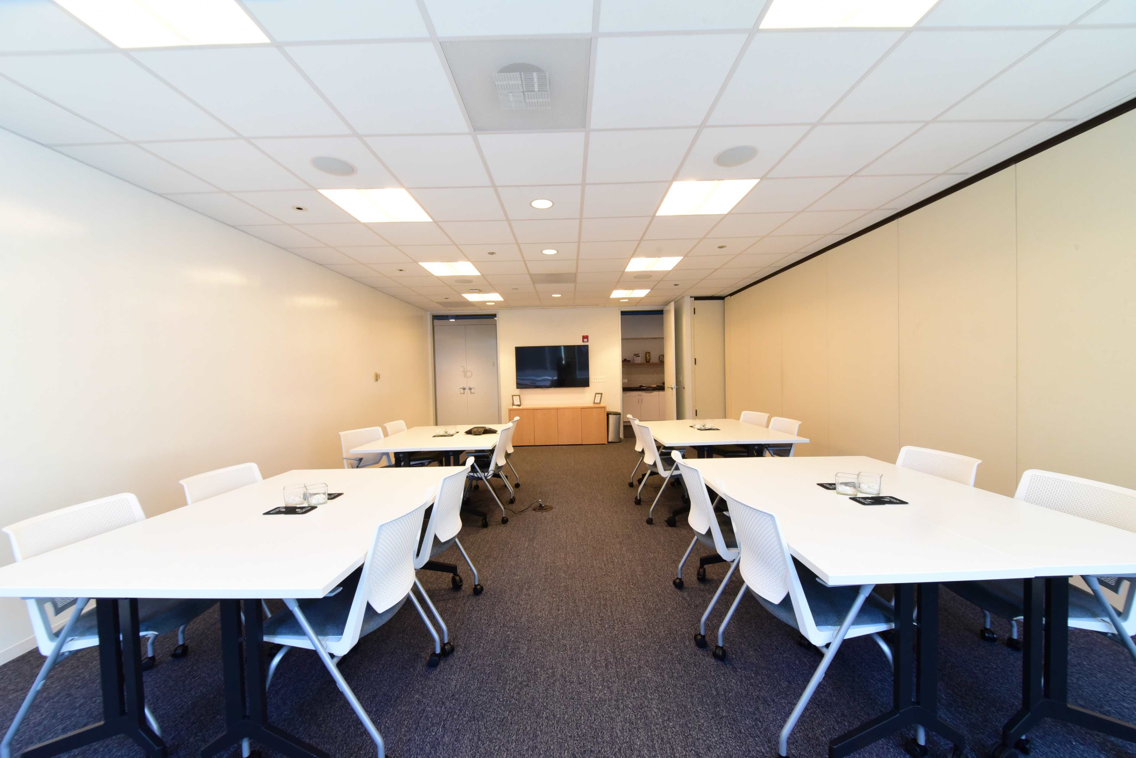 A meeting room with multiple white tables arranged for discussions, white chairs, and a television screen mounted on the wall.