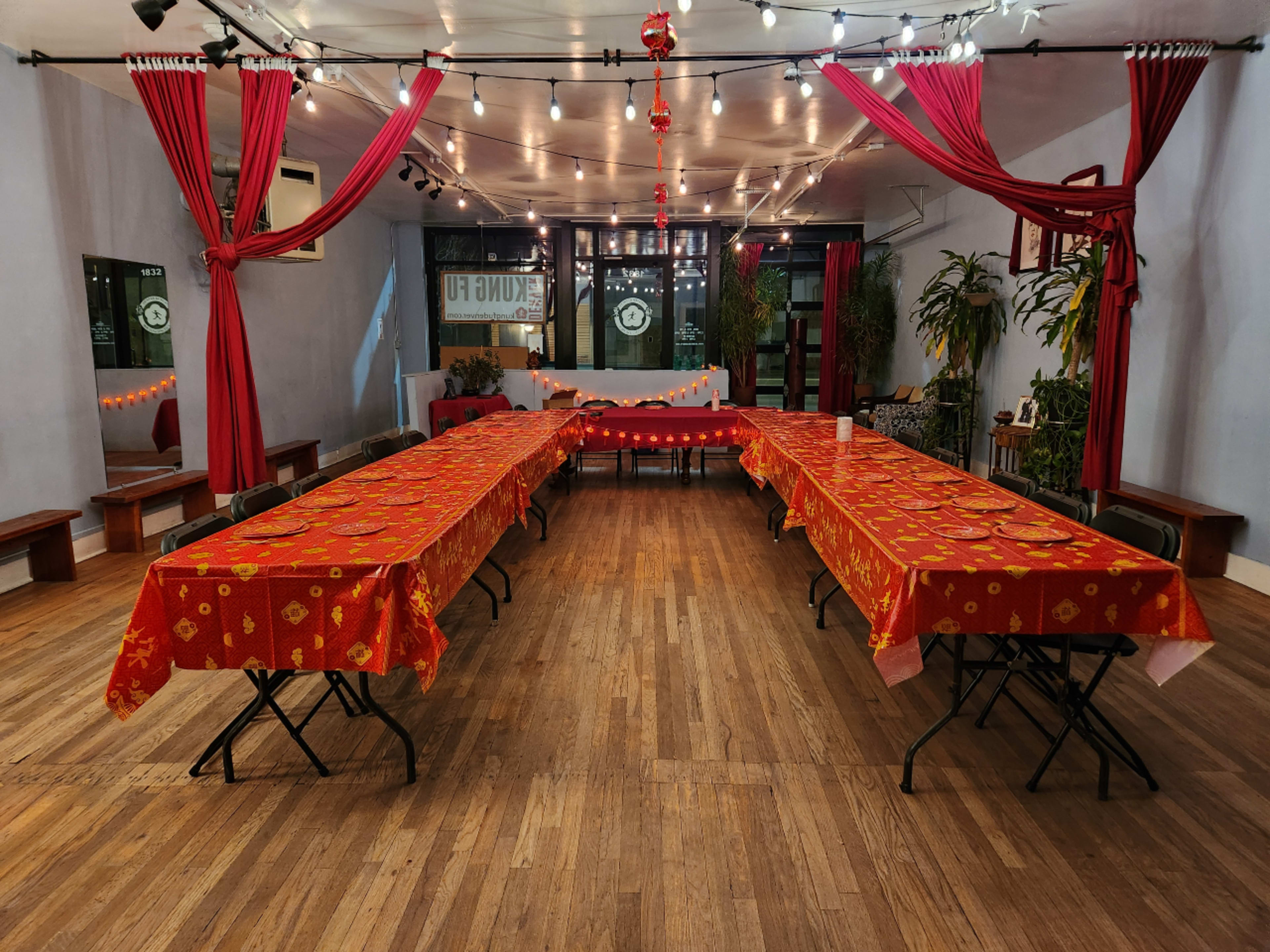 A spacious indoor venue with two long tables covered in red tablecloths, surrounded by plants and decorated with string lights and red curtains.