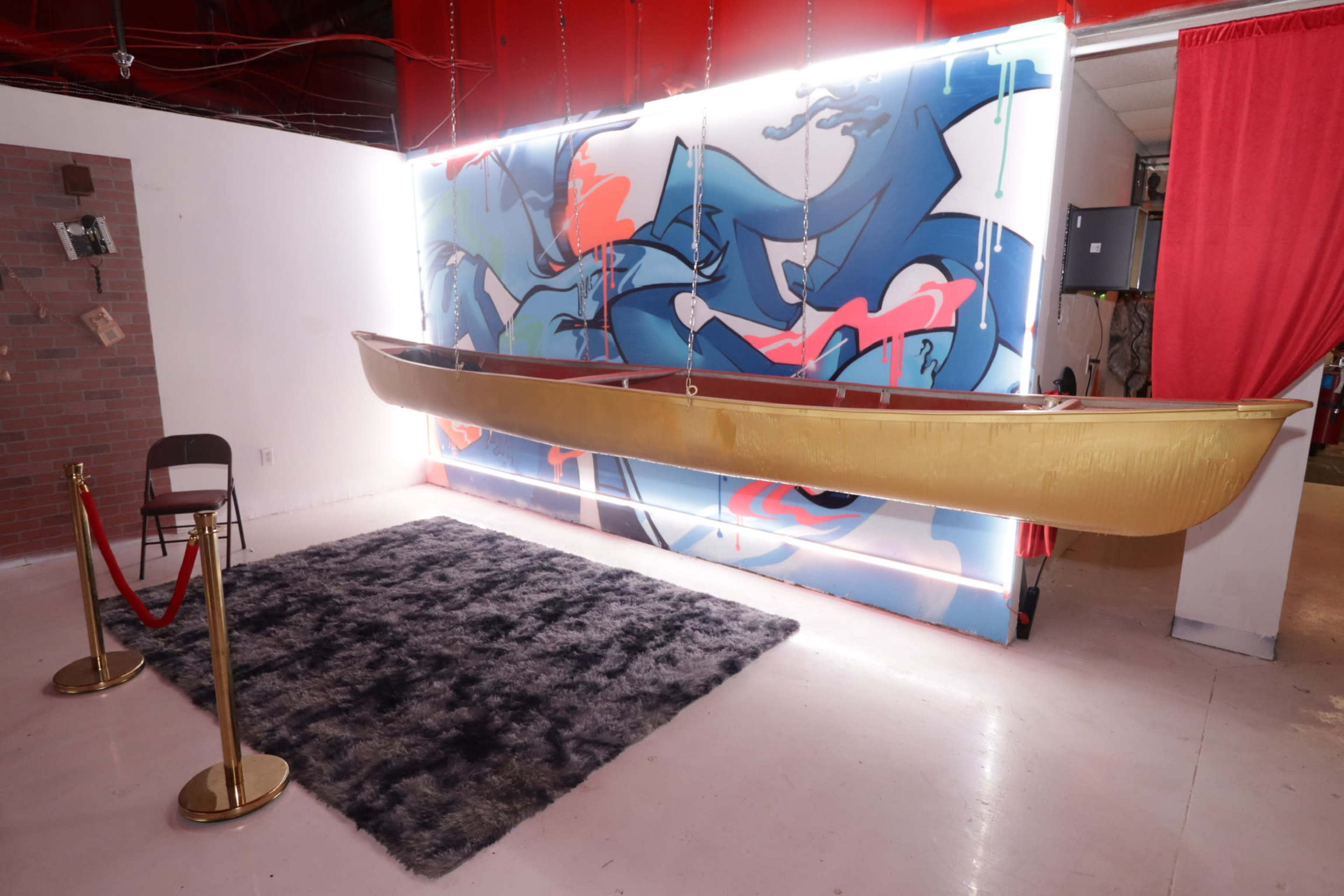 A gold canoe is suspended above a dark rug in front of a colorful mural on a white wall.