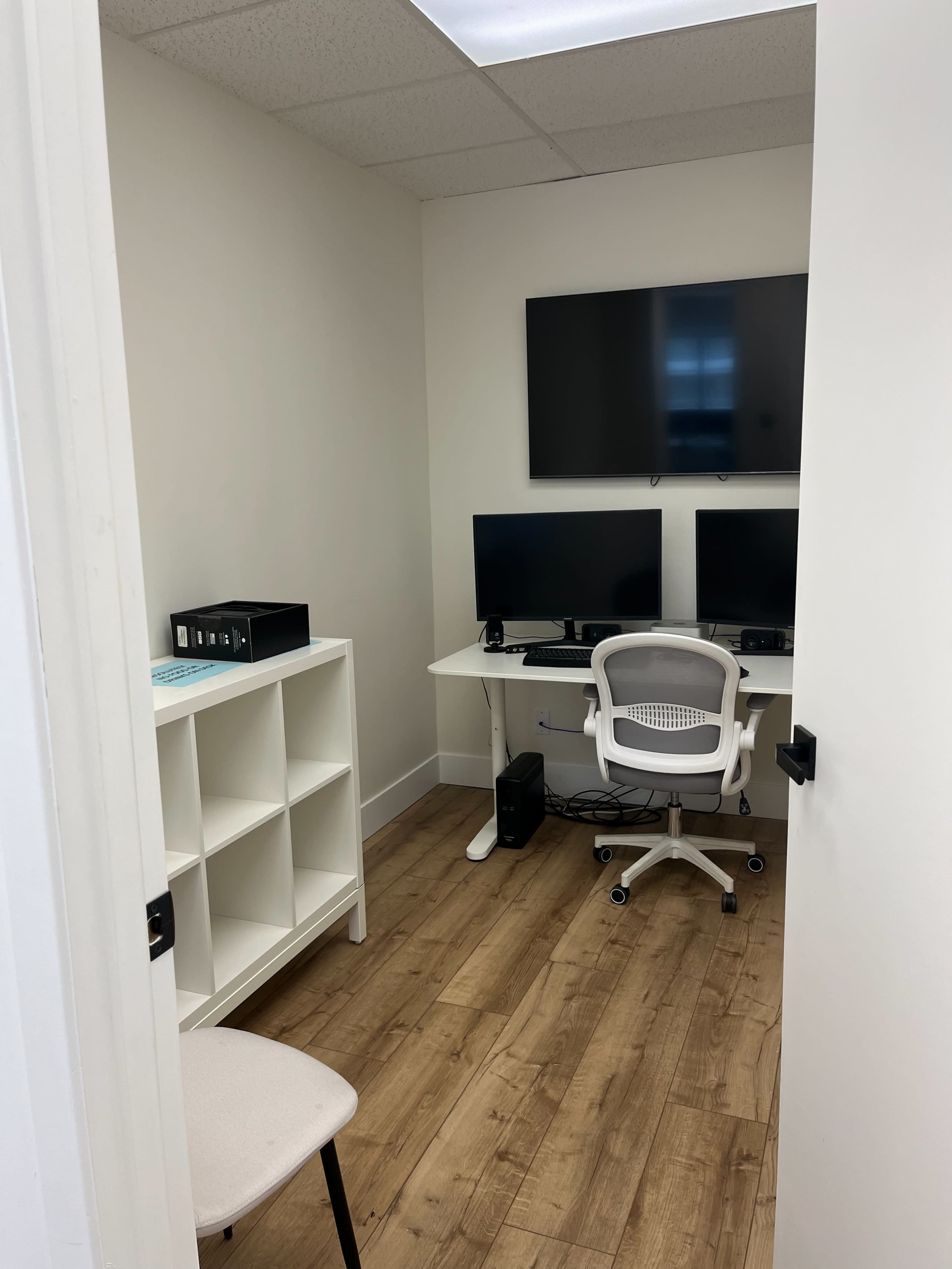 The image shows a small office space with two wall-mounted monitors, a desk with a chair, a storage shelf, and light-colored wooden flooring.
