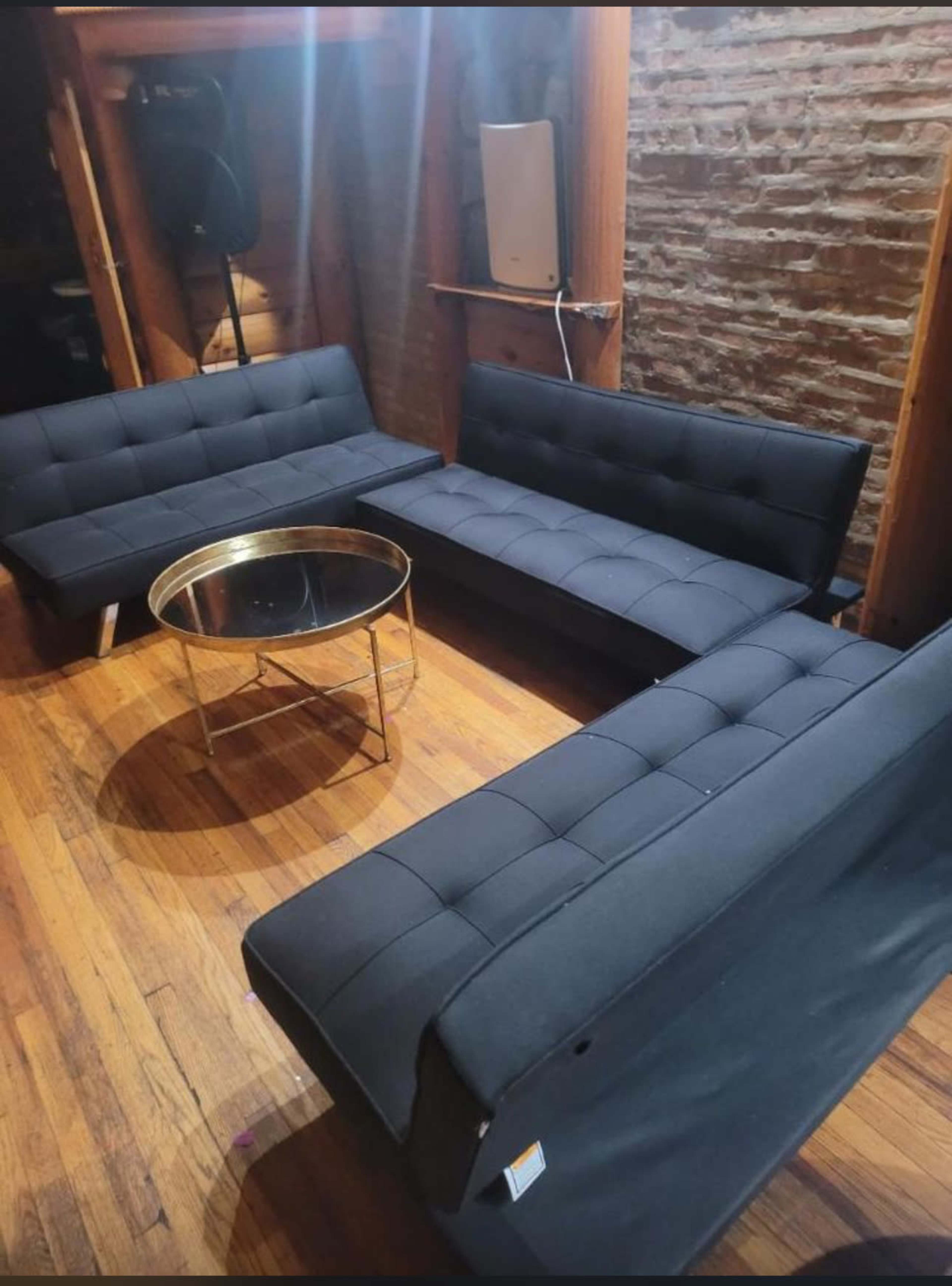 A room with four black futon sofas arranged around a circular gold coffee table on a wooden floor.