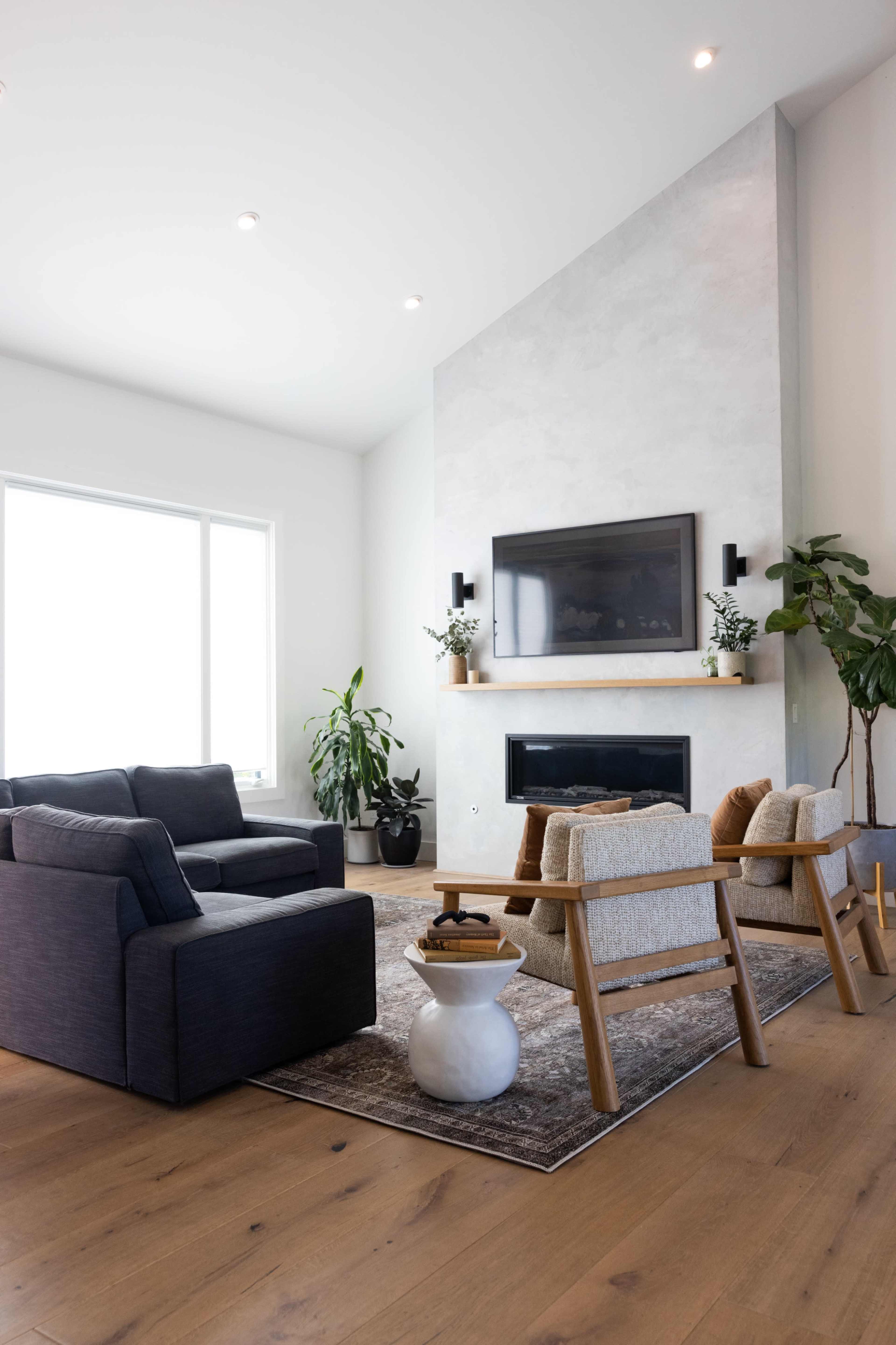 A modern living room with a large window, a fireplace beneath a wall-mounted TV, and various seating options arranged around a central rug.