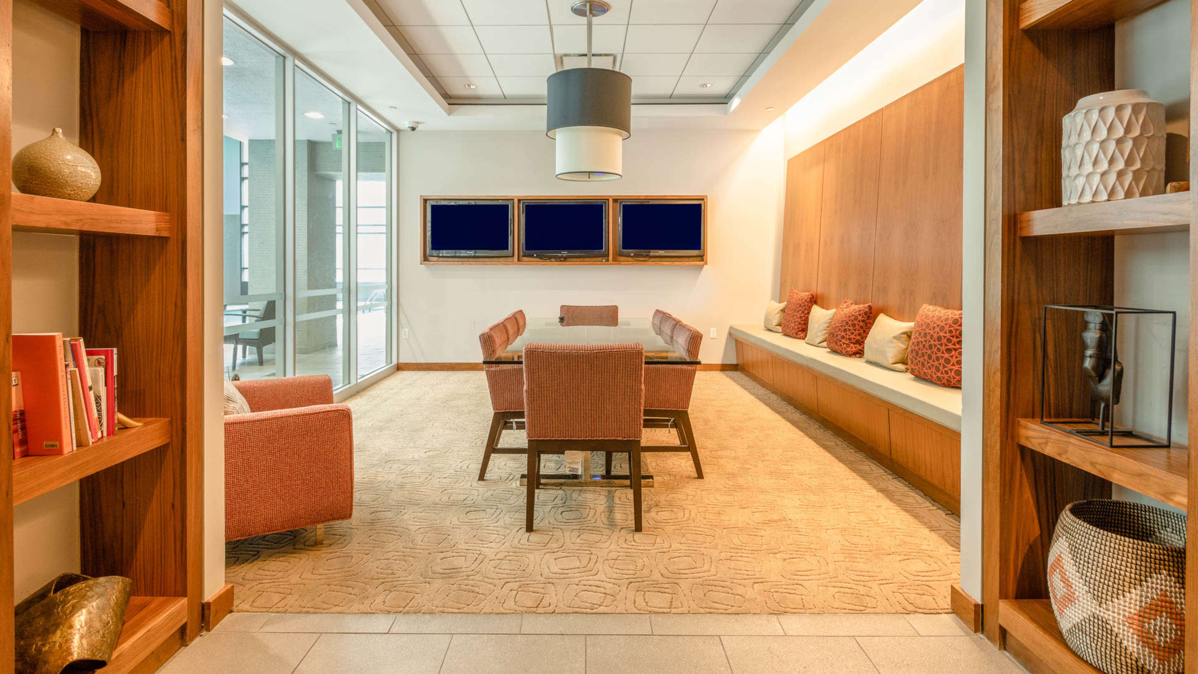 The image shows a modern meeting room with a large table surrounded by chairs, flanked by shelves containing decorative items and books, and featuring wall-mounted screens.