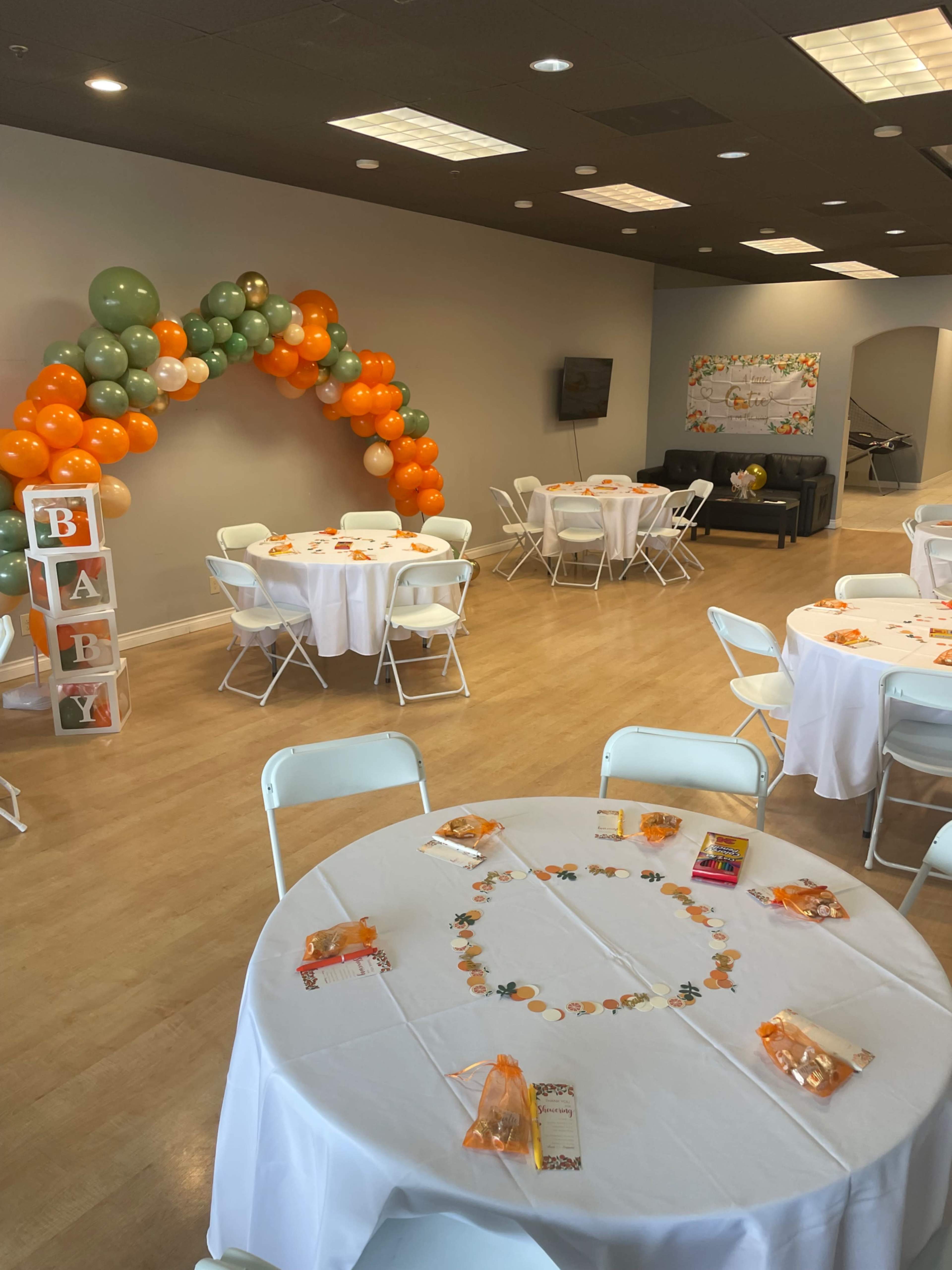 The image shows a decorated event space with round tables, a colorful balloon arch, and a display of orange and green themed decorations for a baby shower.