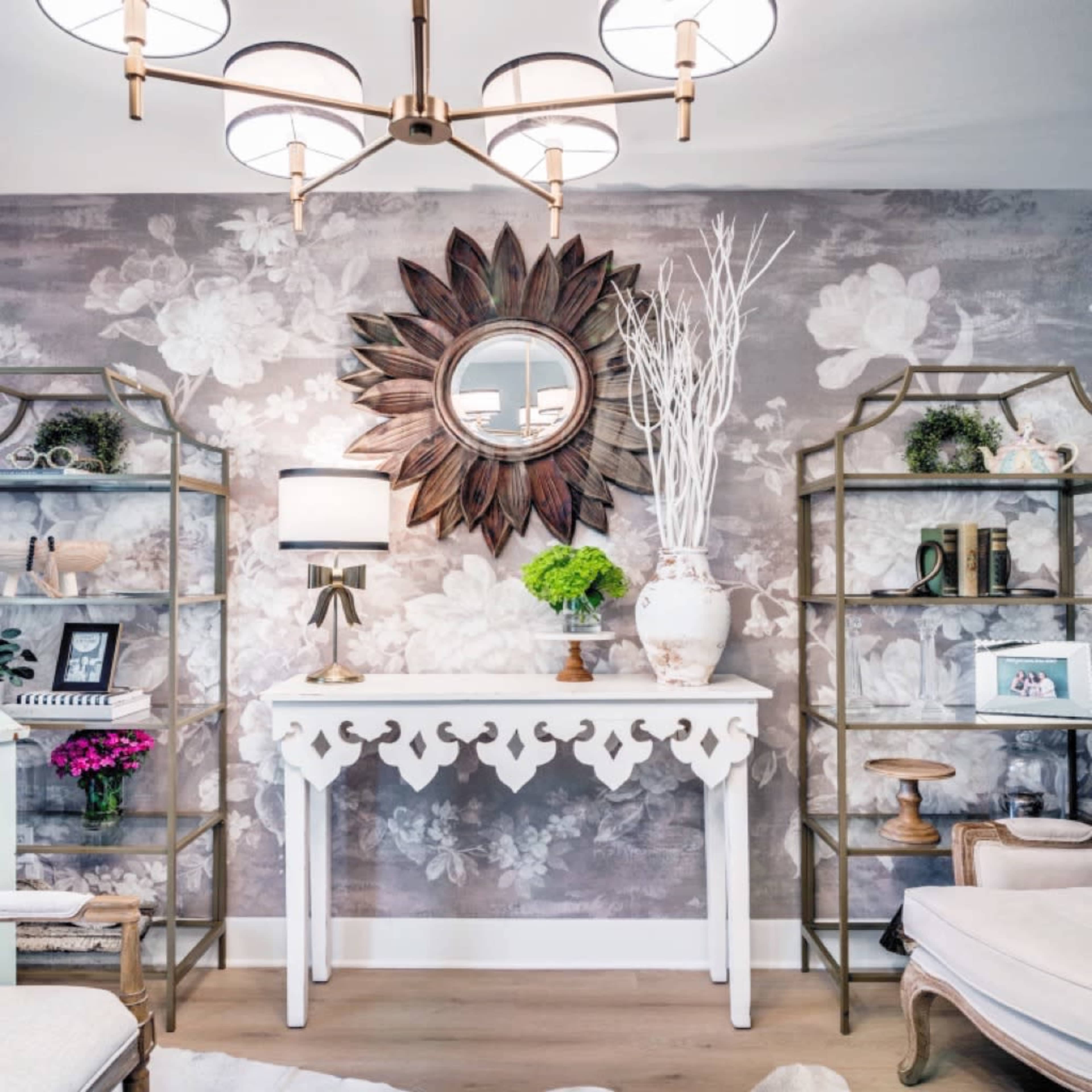 A elegantly decorated room with a floral wallpaper backdrop, a white console table, a round mirror, and two metal shelves holding decorative items and plants.