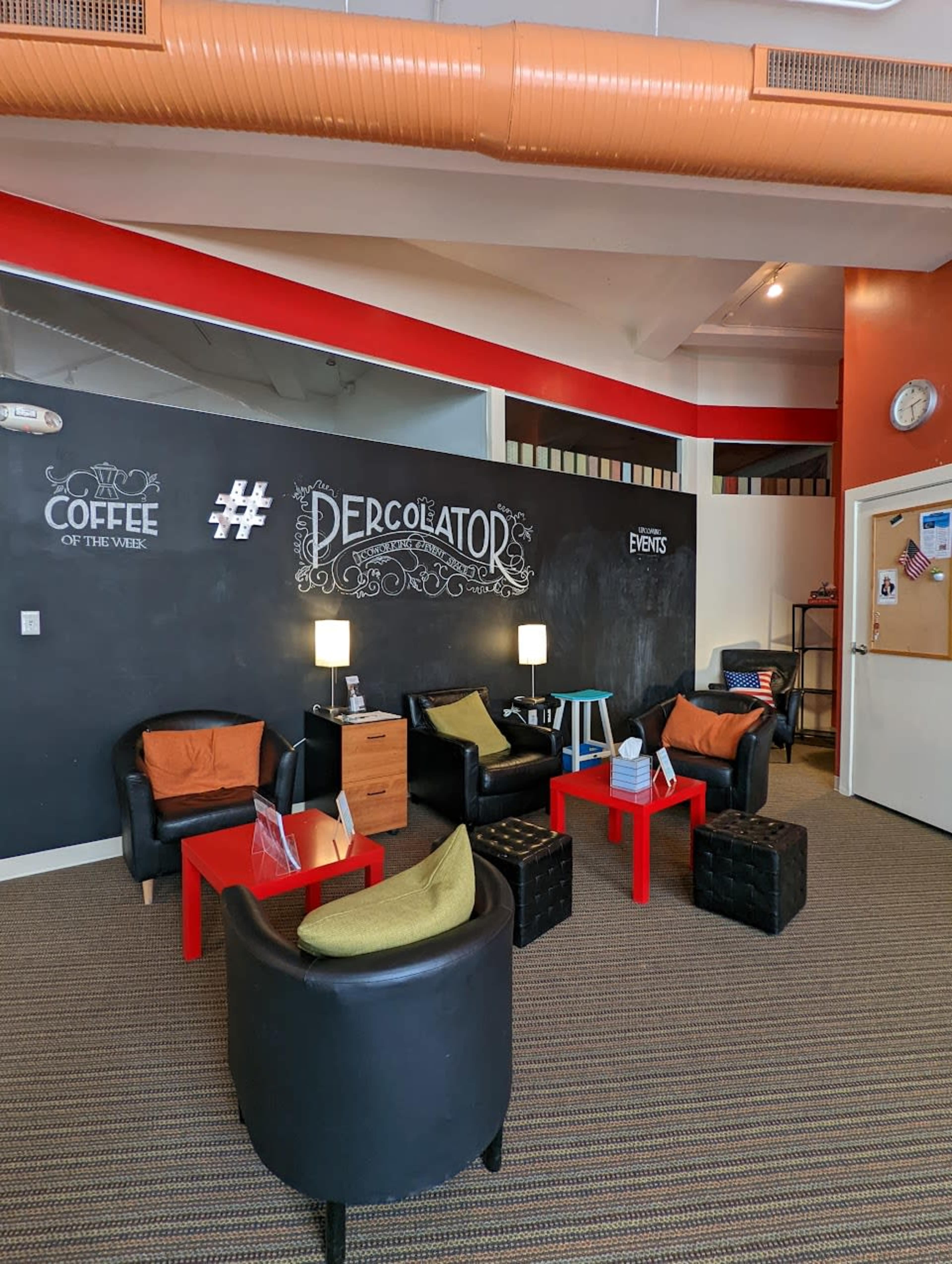The image shows a cozy indoor seating area with black chairs, red side tables, and a large black chalkboard with decorative text, all under a bright ceiling.