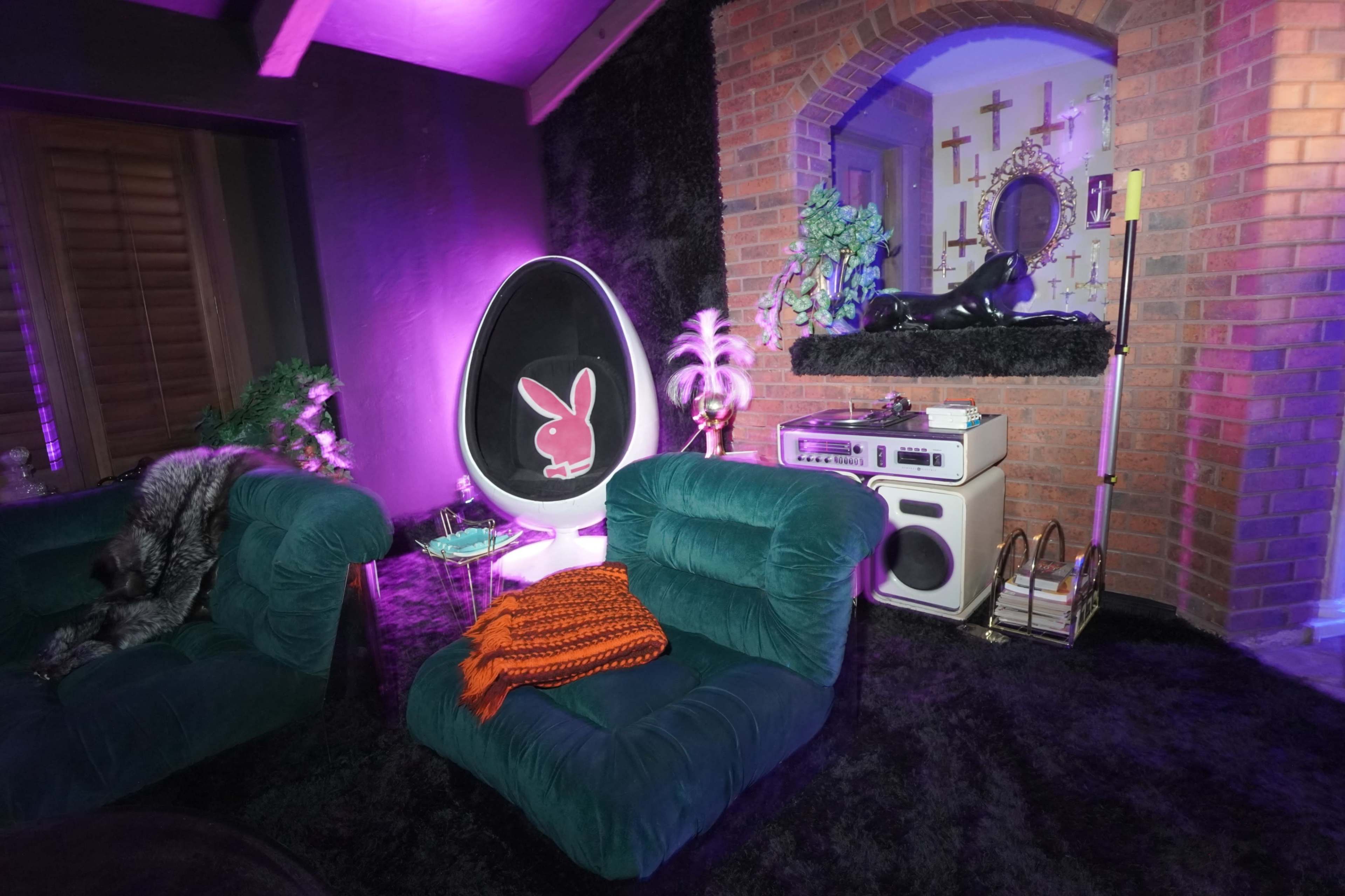 The room features two green velvet chairs, a circular black and pink chair, a vintage boombox, and a decorated wall with bricks and various decorative items in moody lighting.