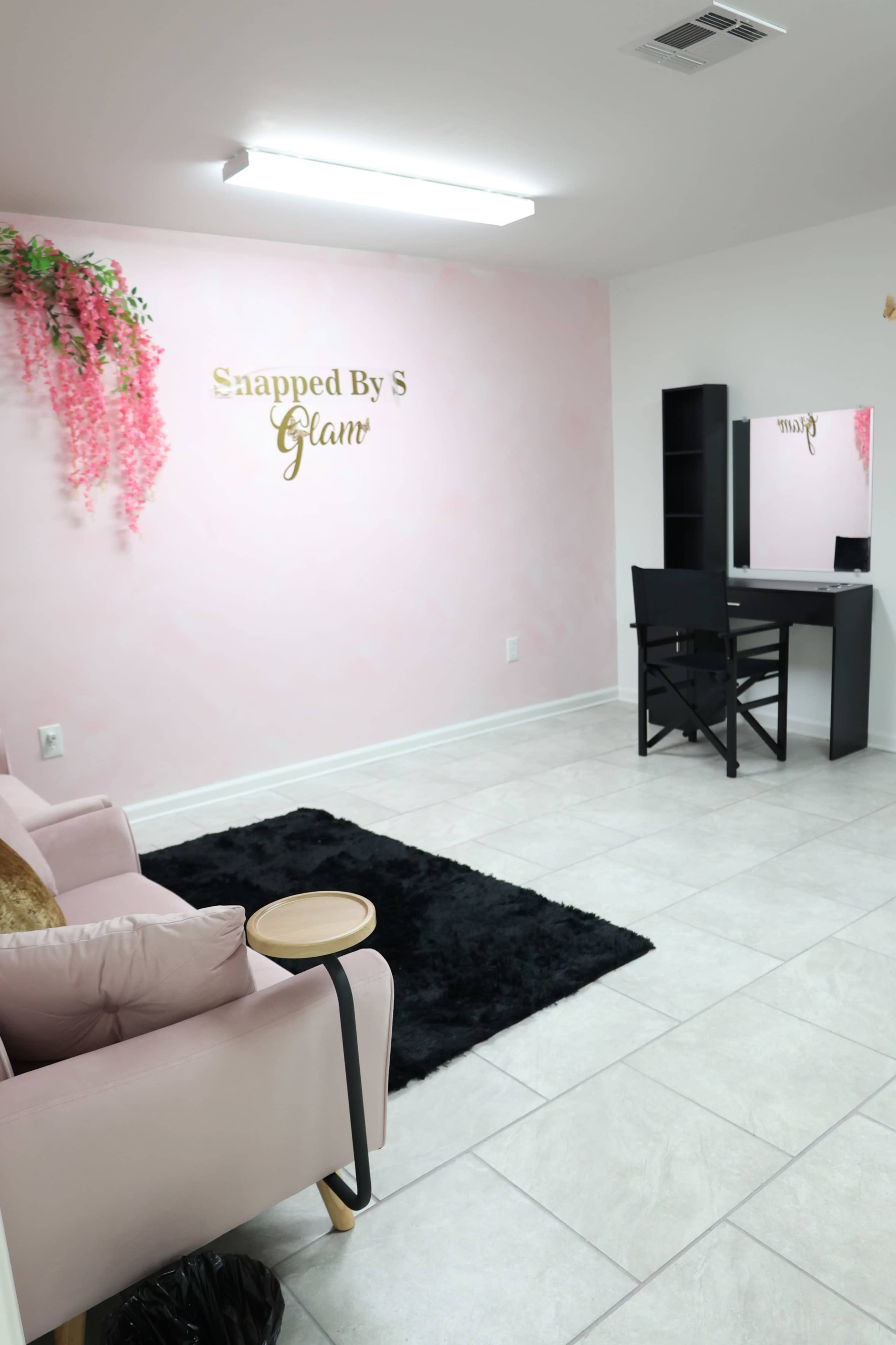 The image shows a light pink room with a plush sofa, a small black rug, a vanity mirror, and a desk against a wall adorned with artificial pink flowers.