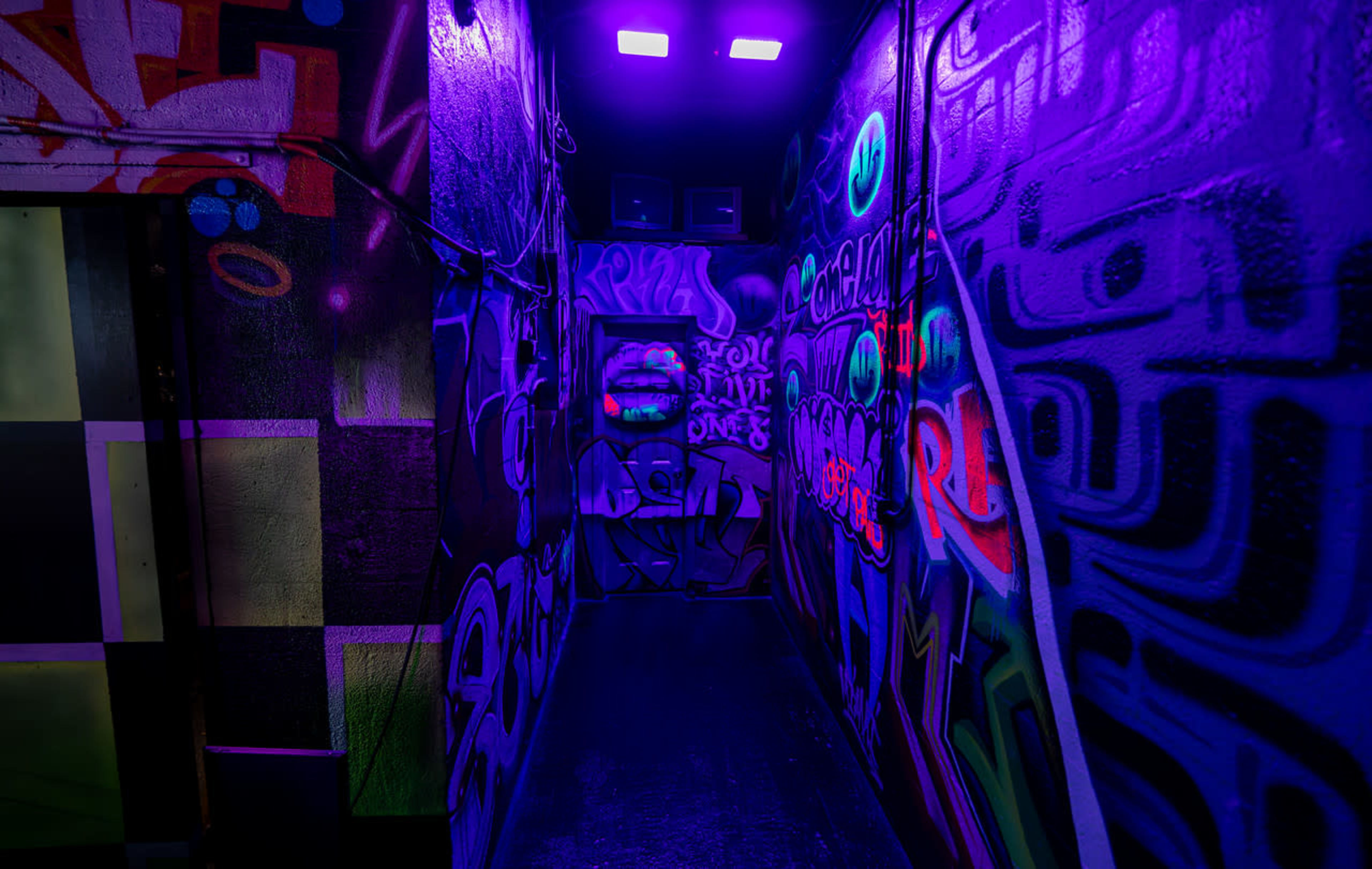 A narrow hallway illuminated by purple lights, with colorful graffiti covering the walls.