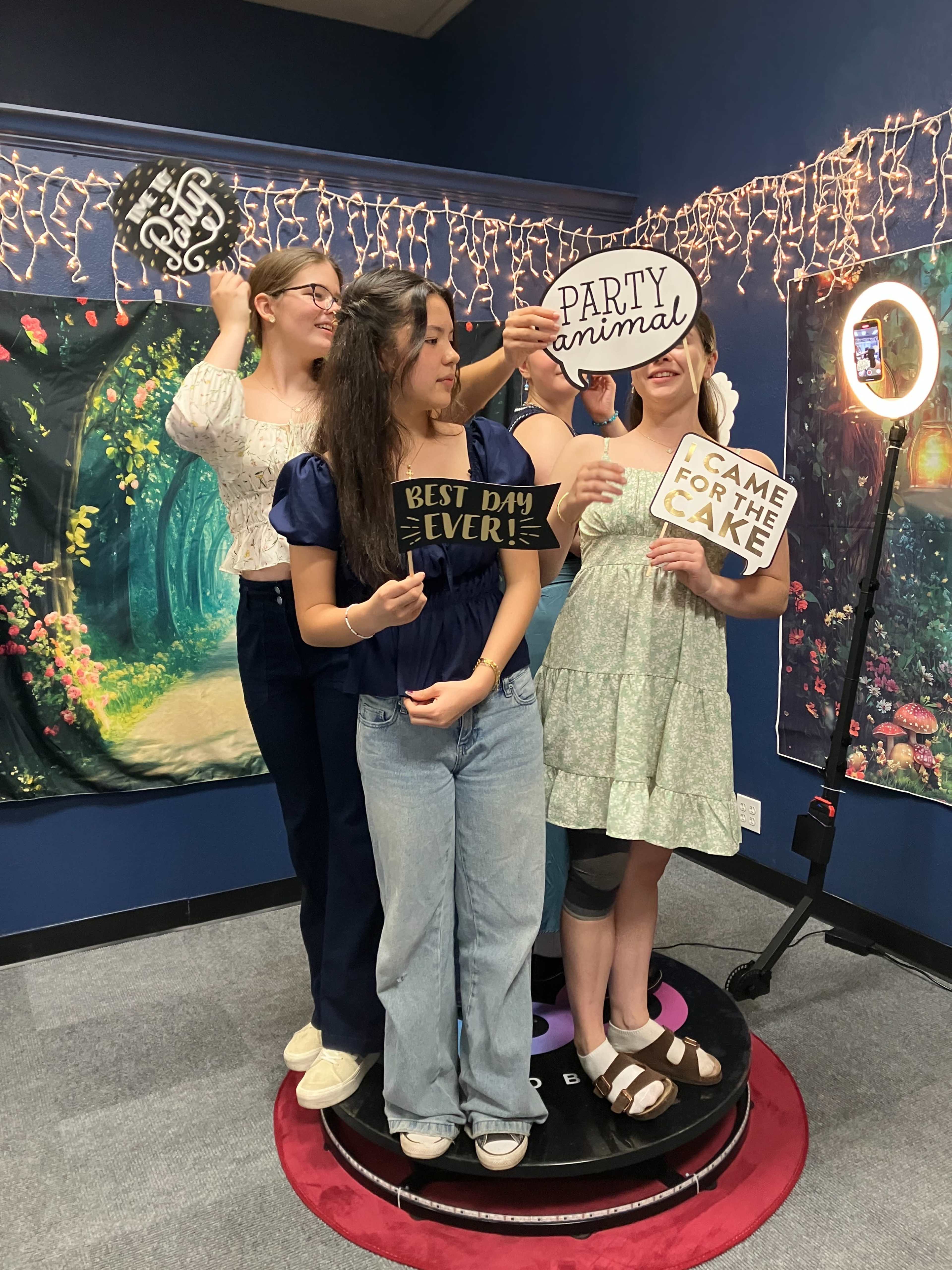 Four young women are standing together on a small platform, each holding a sign with various phrases while a ring light illuminates the scene.