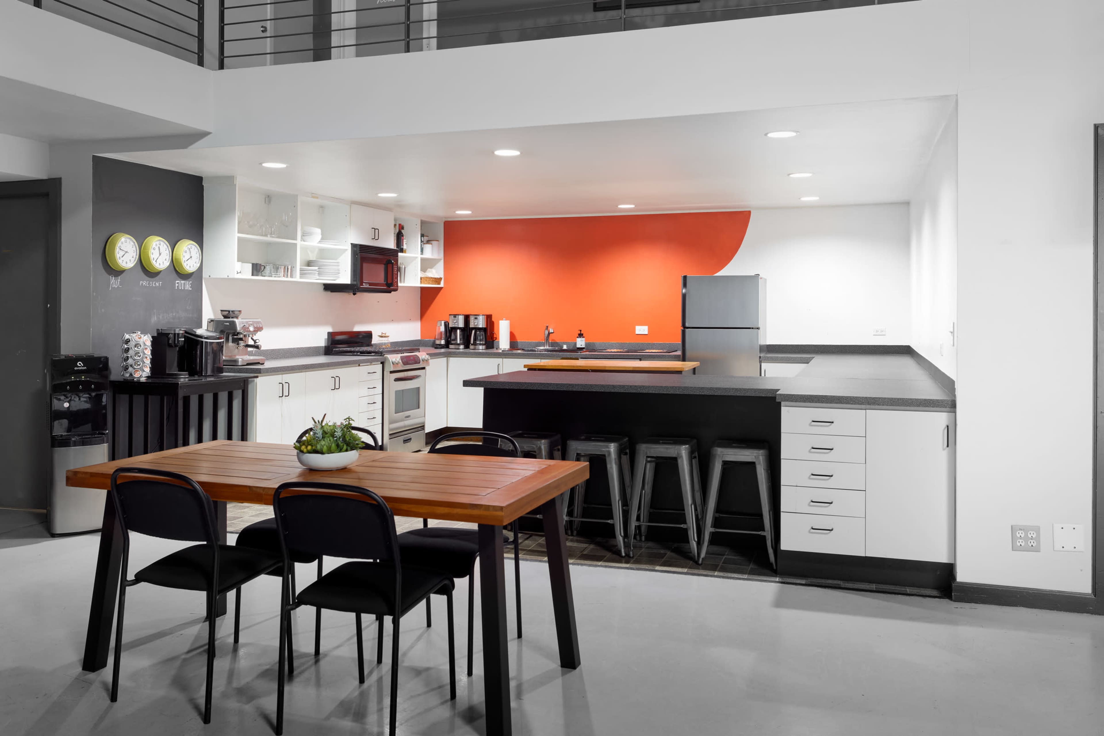 The image shows a modern kitchen area featuring a wooden dining table with black chairs, a countertop with bar stools, and an orange accent wall.