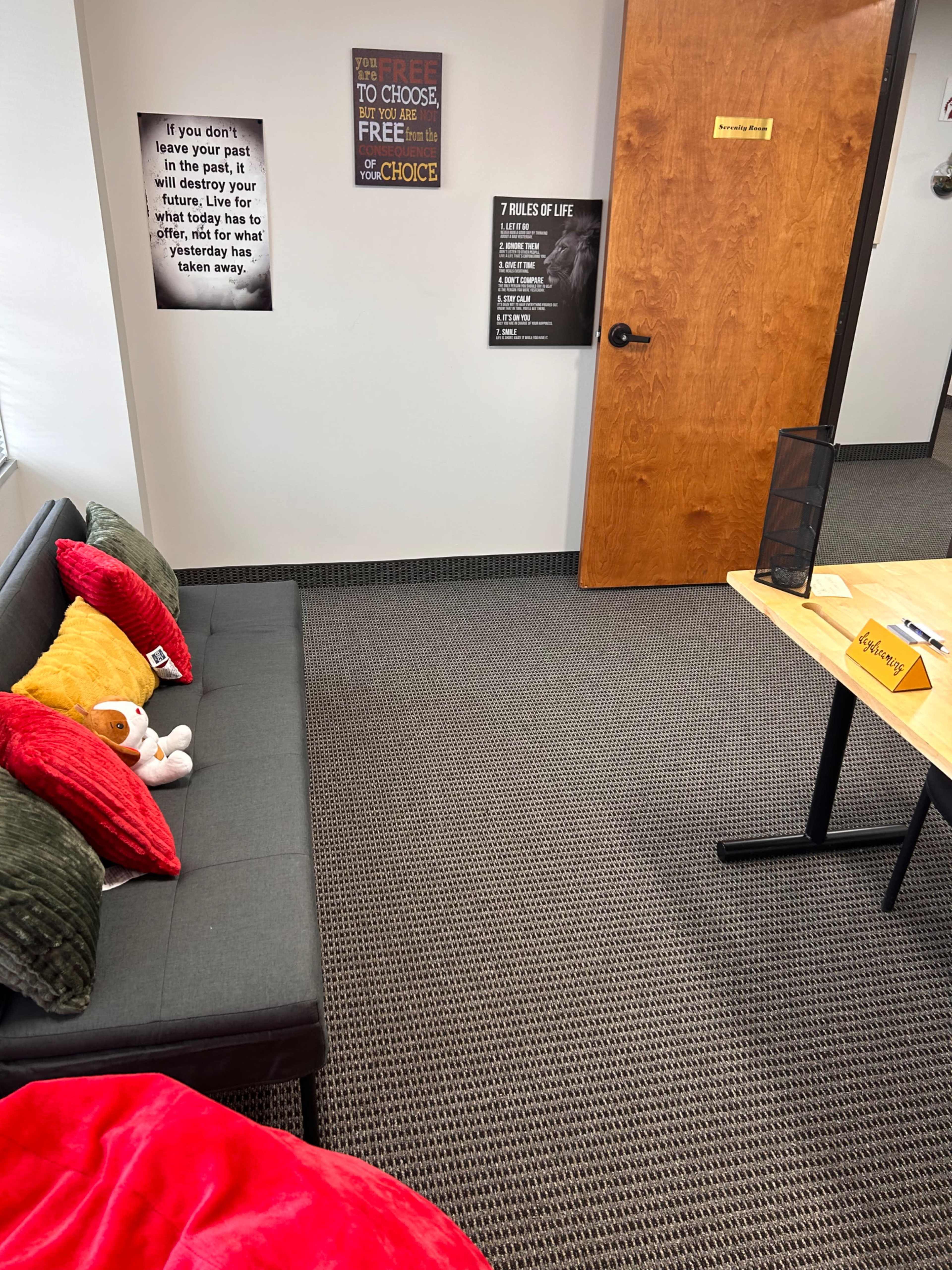 The image shows a cozy office space with a gray sofa covered in pillows, a red blanket, a small table with a sign, and motivational posters on the walls.