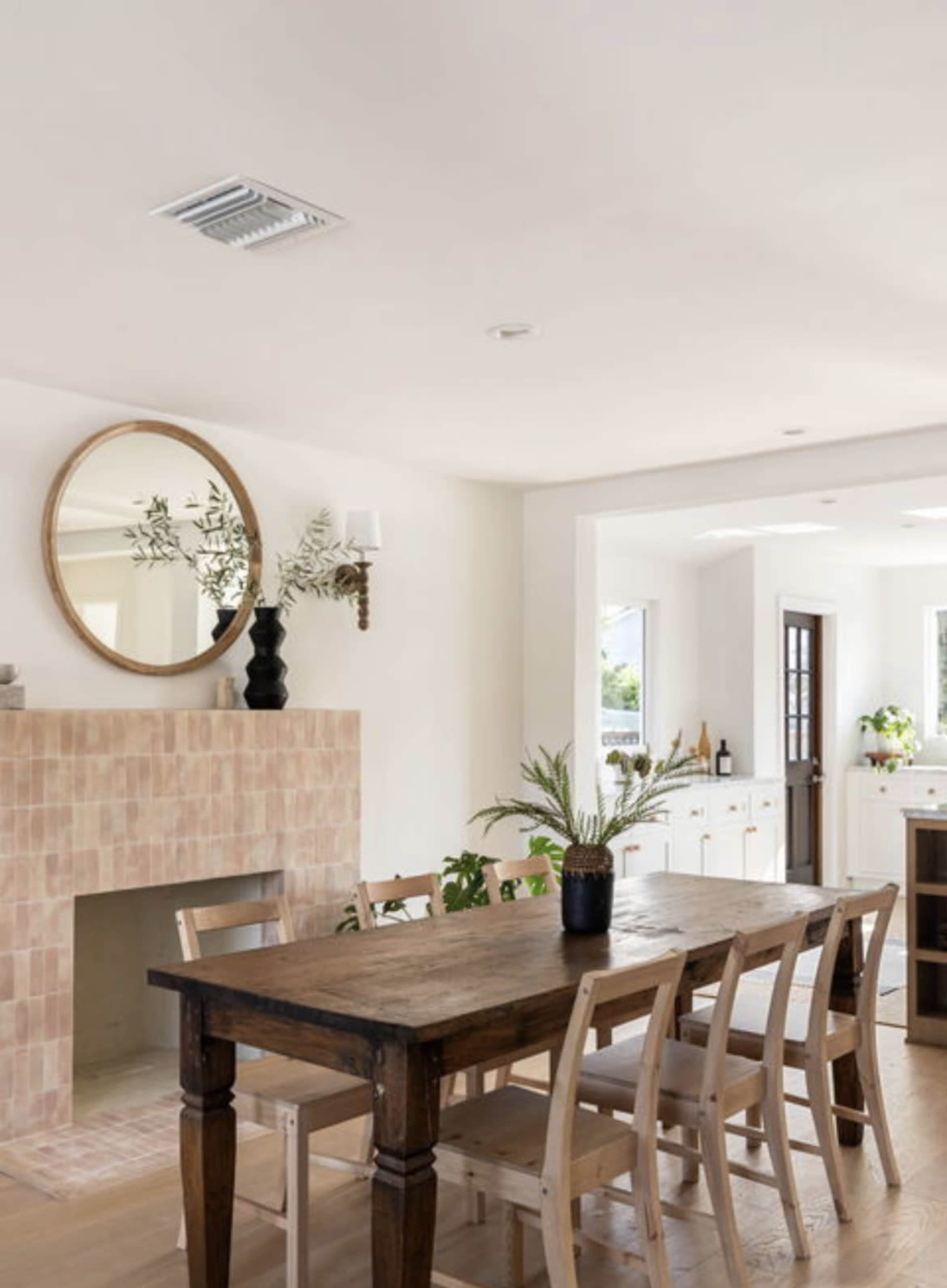 A wooden dining table with chairs is positioned near a fireplace underneath a large round mirror in a bright, modern interior.