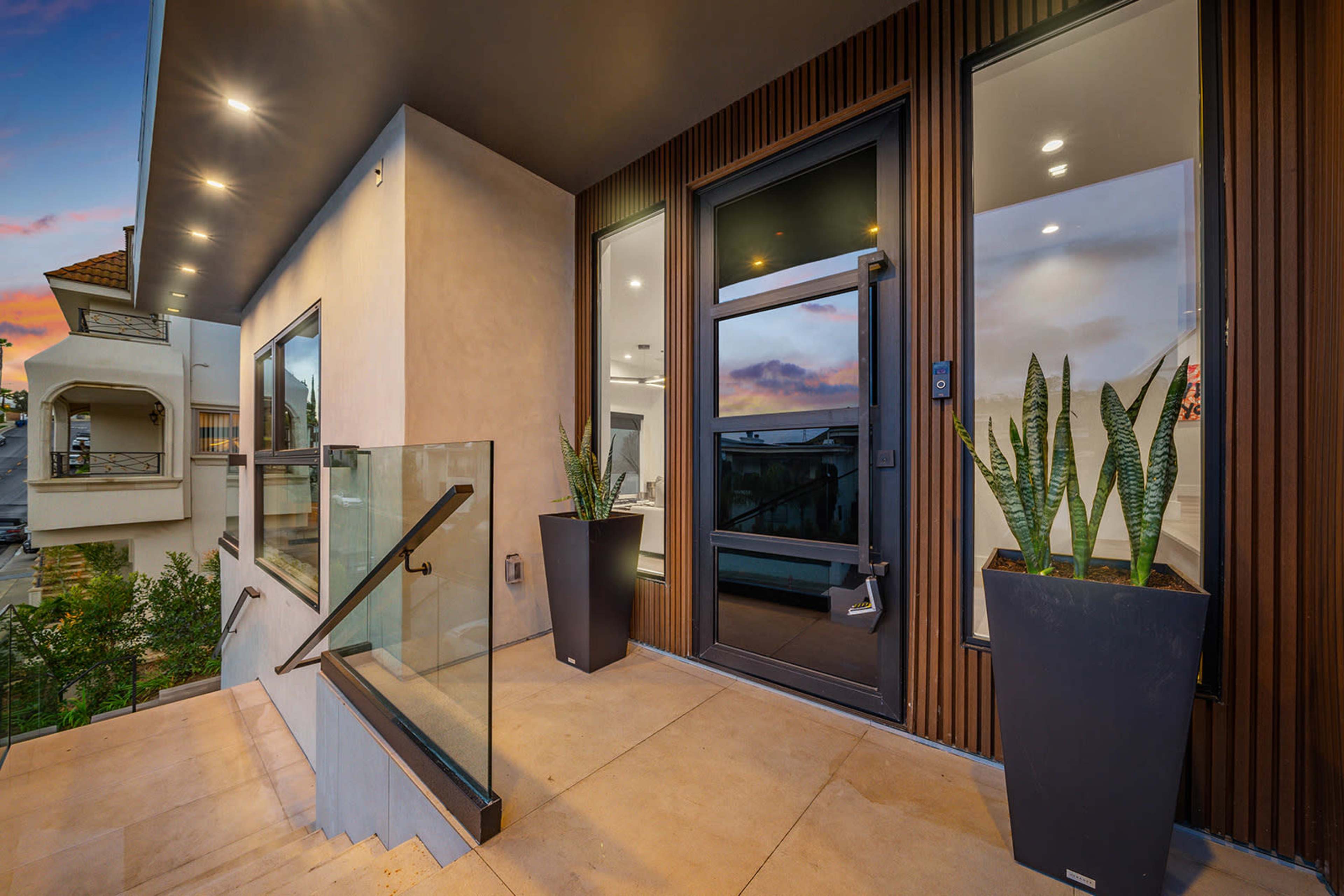 The image shows a modern entrance with glass railings, tall planters, and large windows, leading to a door framed by horizontal wooden panels.