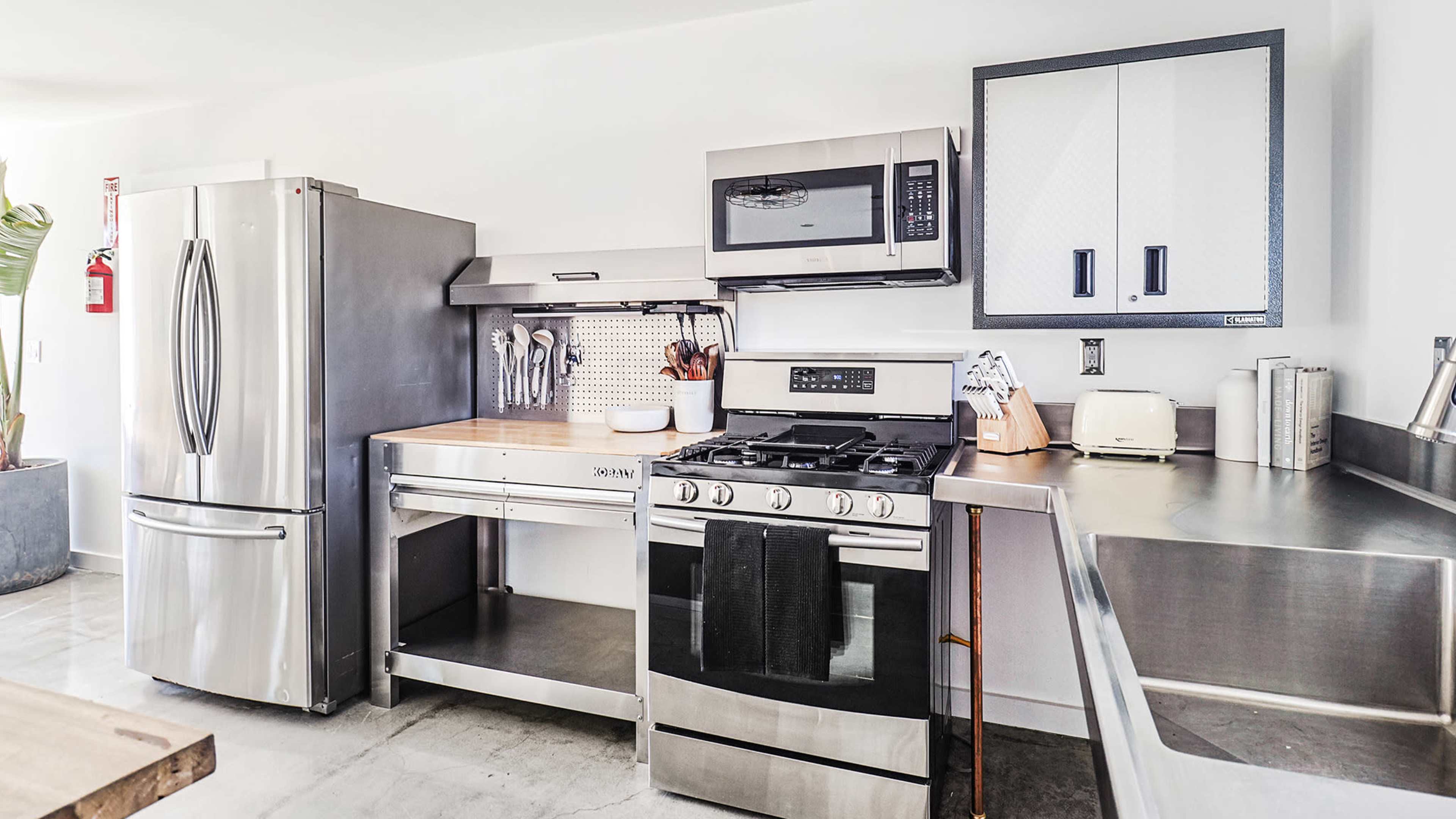 A modern kitchen equipped with stainless steel appliances, including a refrigerator, microwave, and gas stove, set against a minimalist countertop and cabinetry.