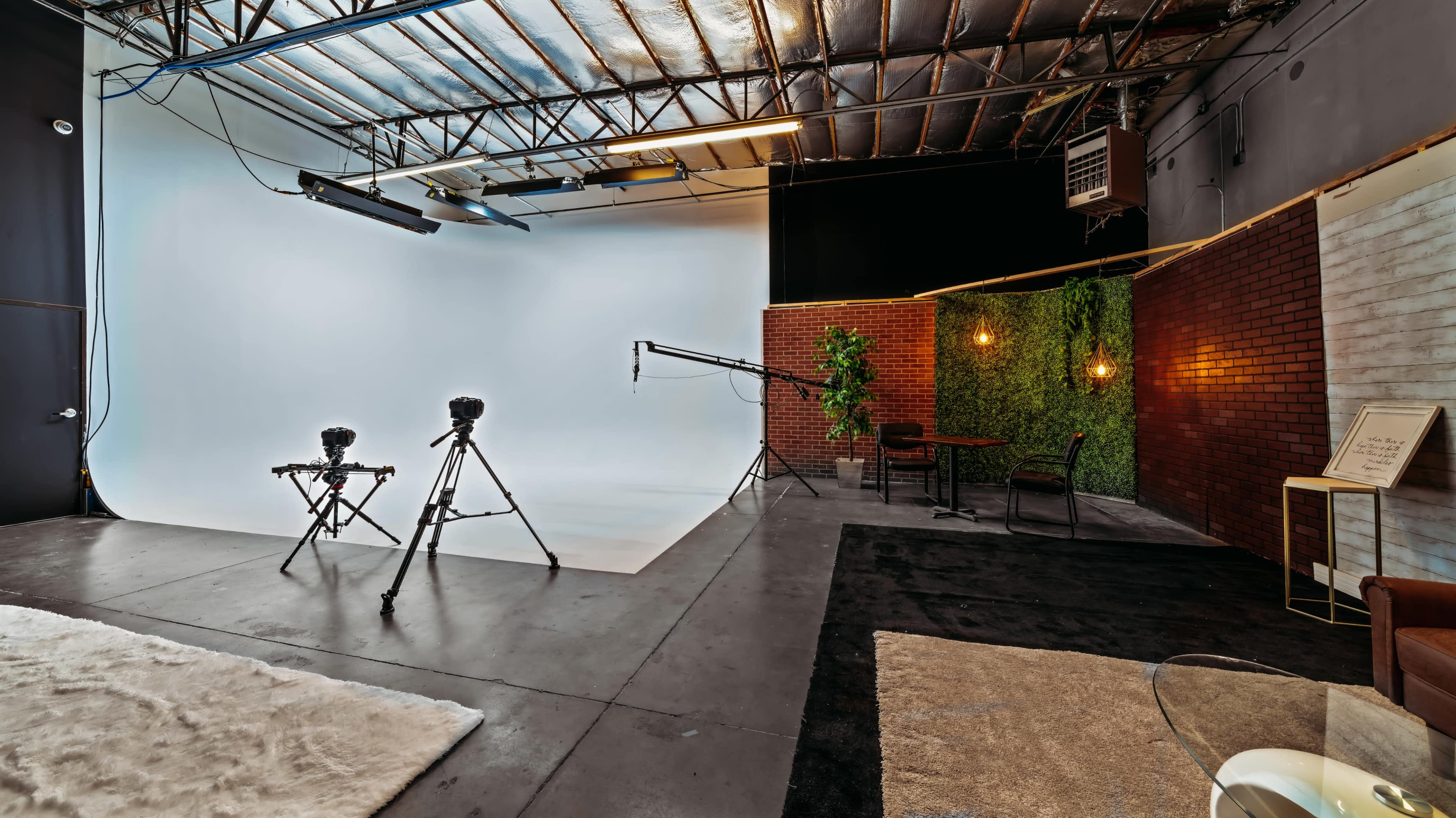 A photography studio featuring two tripods, a light setup, a green wall, and seating arrangements against a backdrop of white and brick walls.