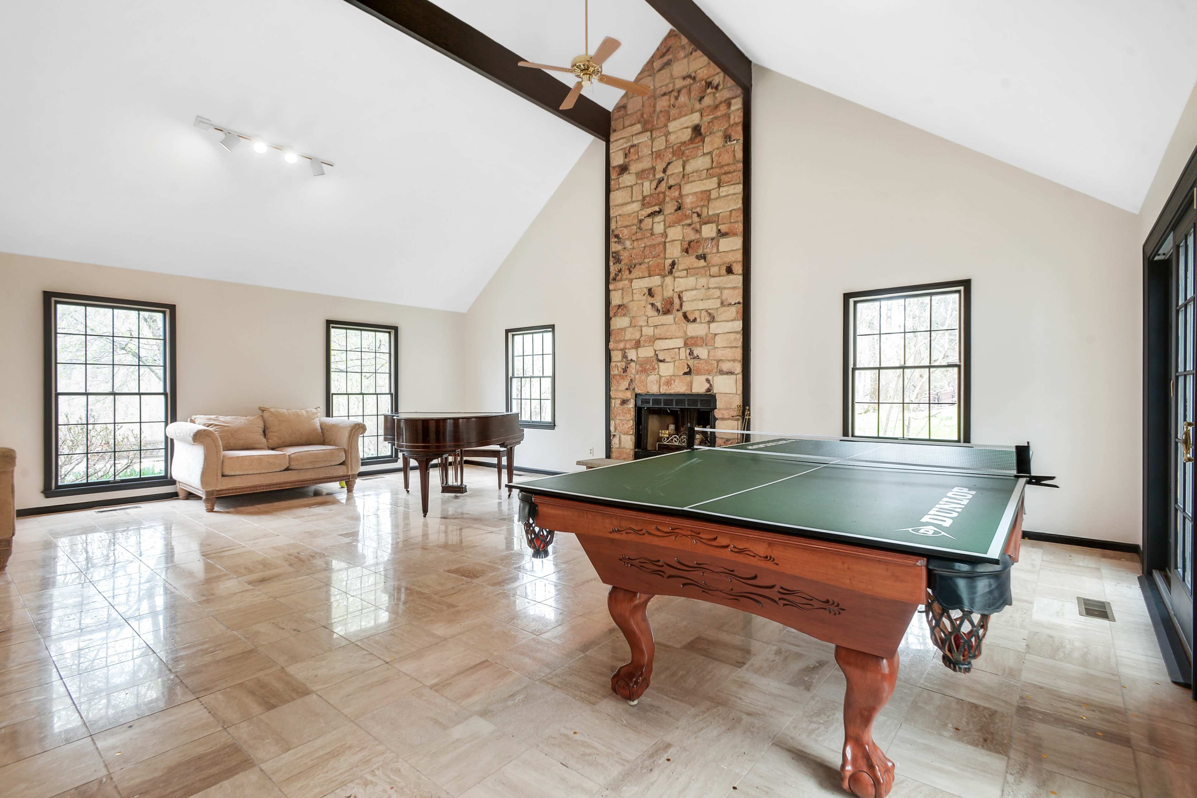 The room features a pool table alongside a piano, with large windows providing natural light and a stone fireplace as a focal point.