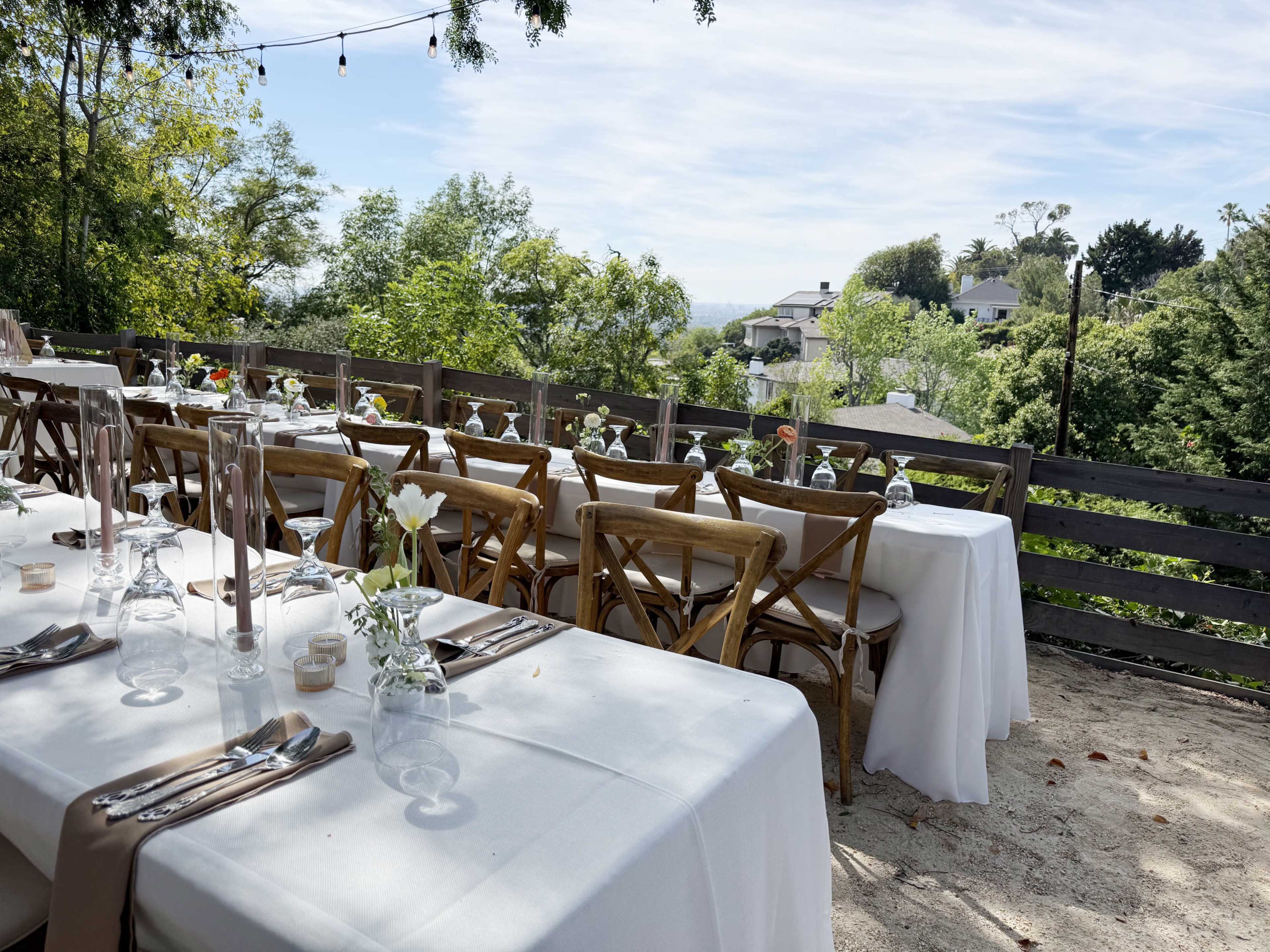 A dining setup with white tablecloths and wooden chairs is arranged outdoors with a view of trees and distant houses.