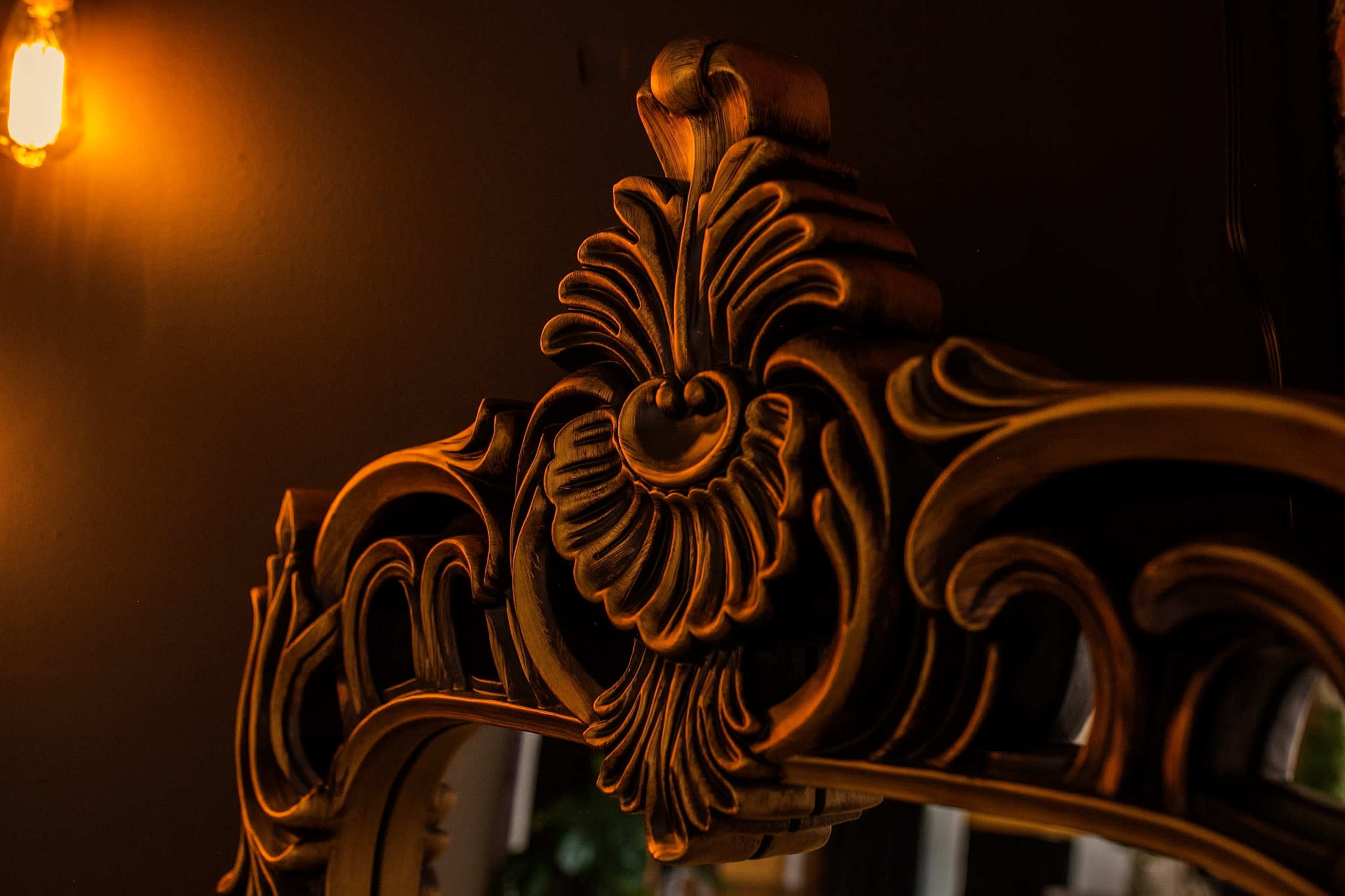 The image shows a close-up of an ornate, gold-colored mirror frame with intricate floral designs and a light bulb in the background.
