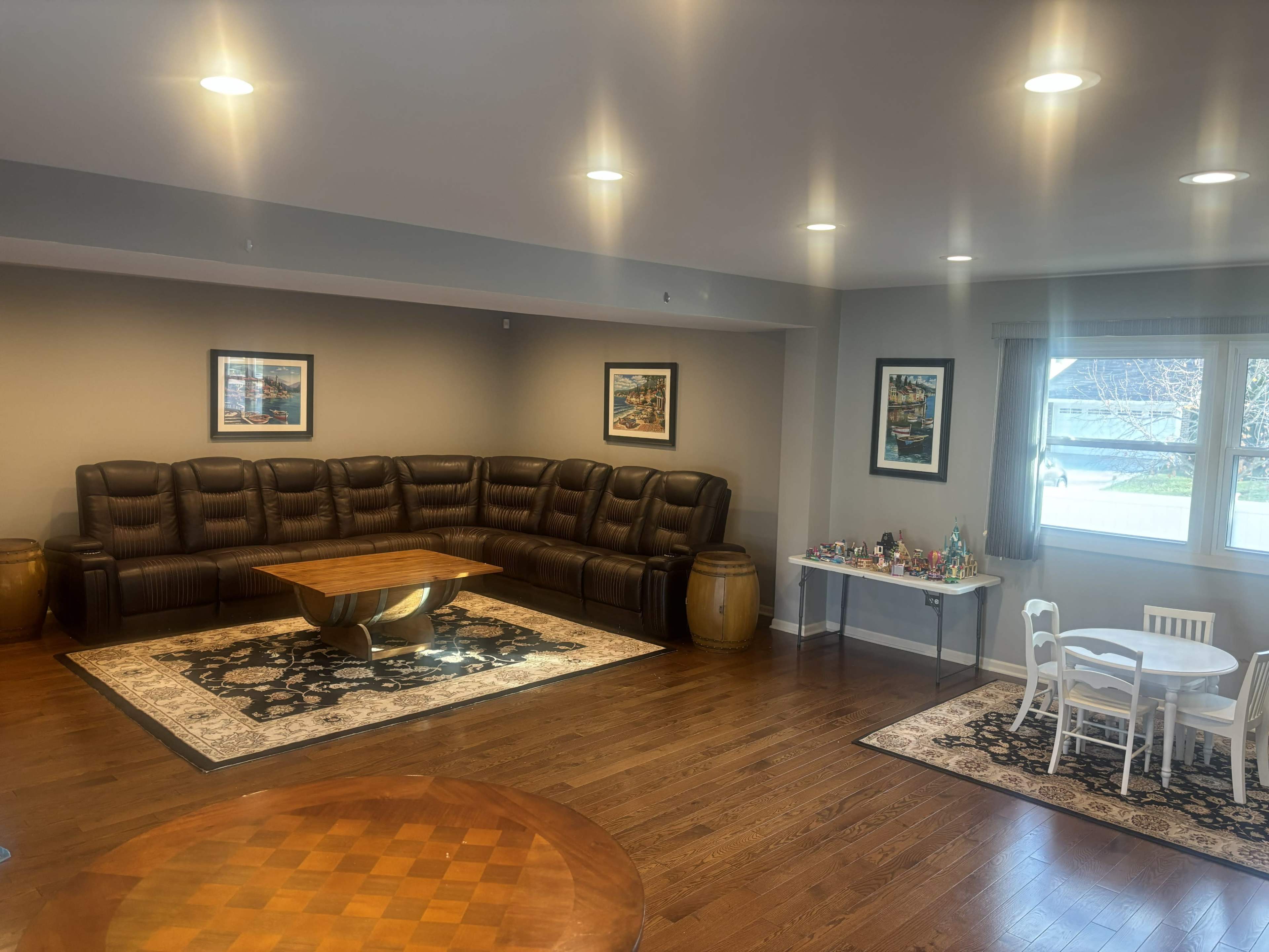 The image shows a spacious living room featuring a large sectional sofa, a round wooden coffee table, decorative area rugs, and a small dining table set with chairs.