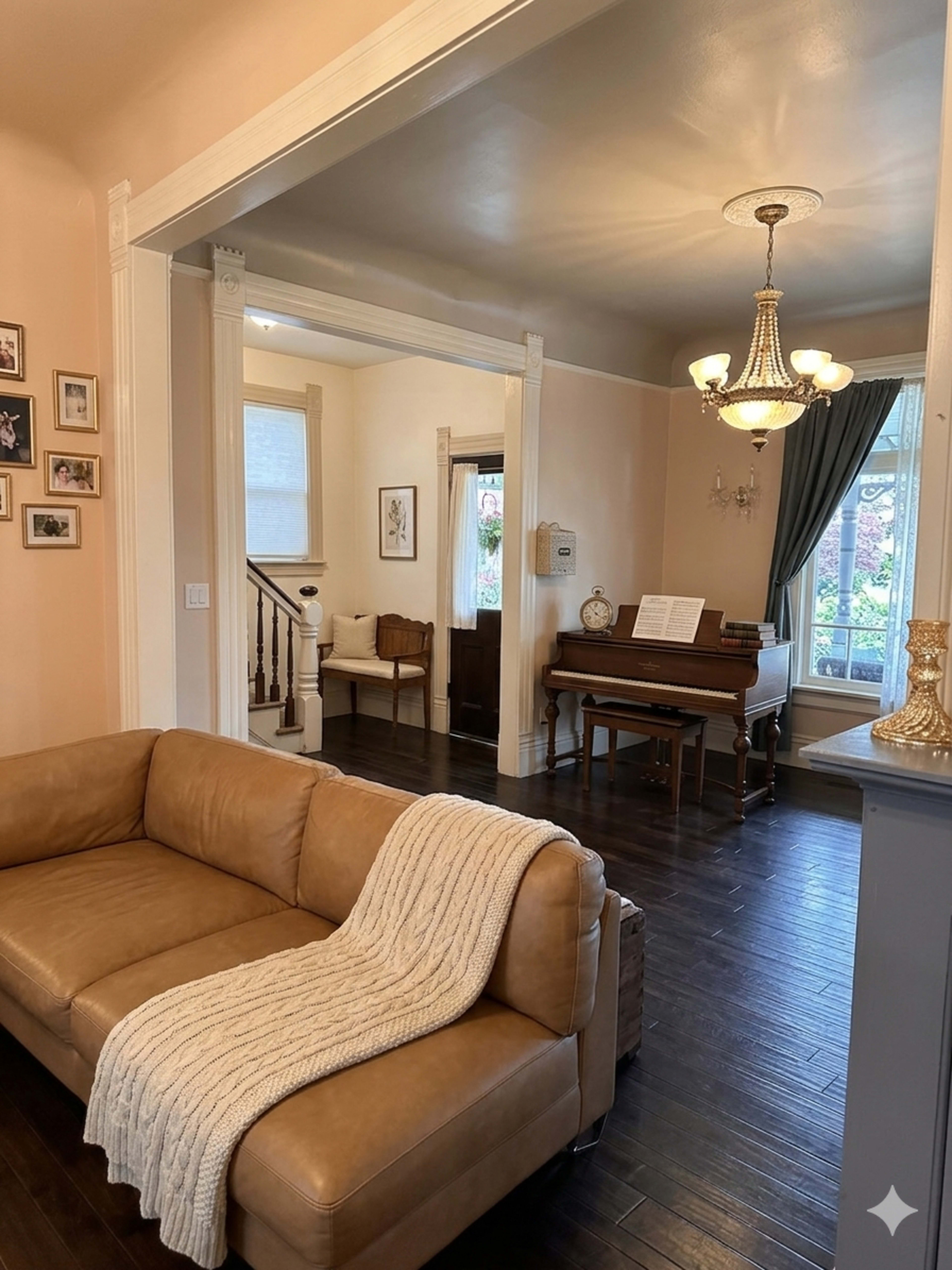 The image shows a light-colored living room with a sectional sofa, a piano, and a chandelier, leading to a hallway and staircase.