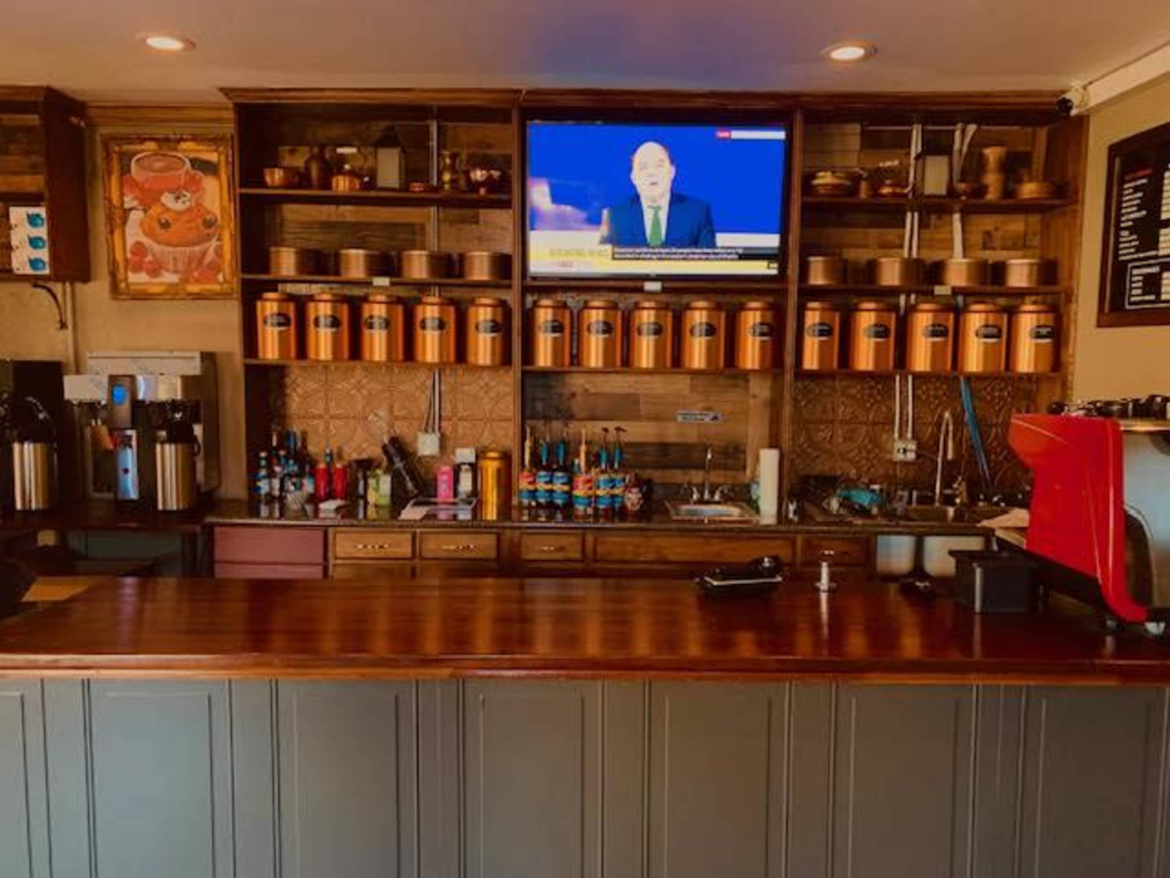 The image shows a coffee shop counter with storage shelves, a television mounted above displaying news, and various coffee equipment and supplies arranged neatly.