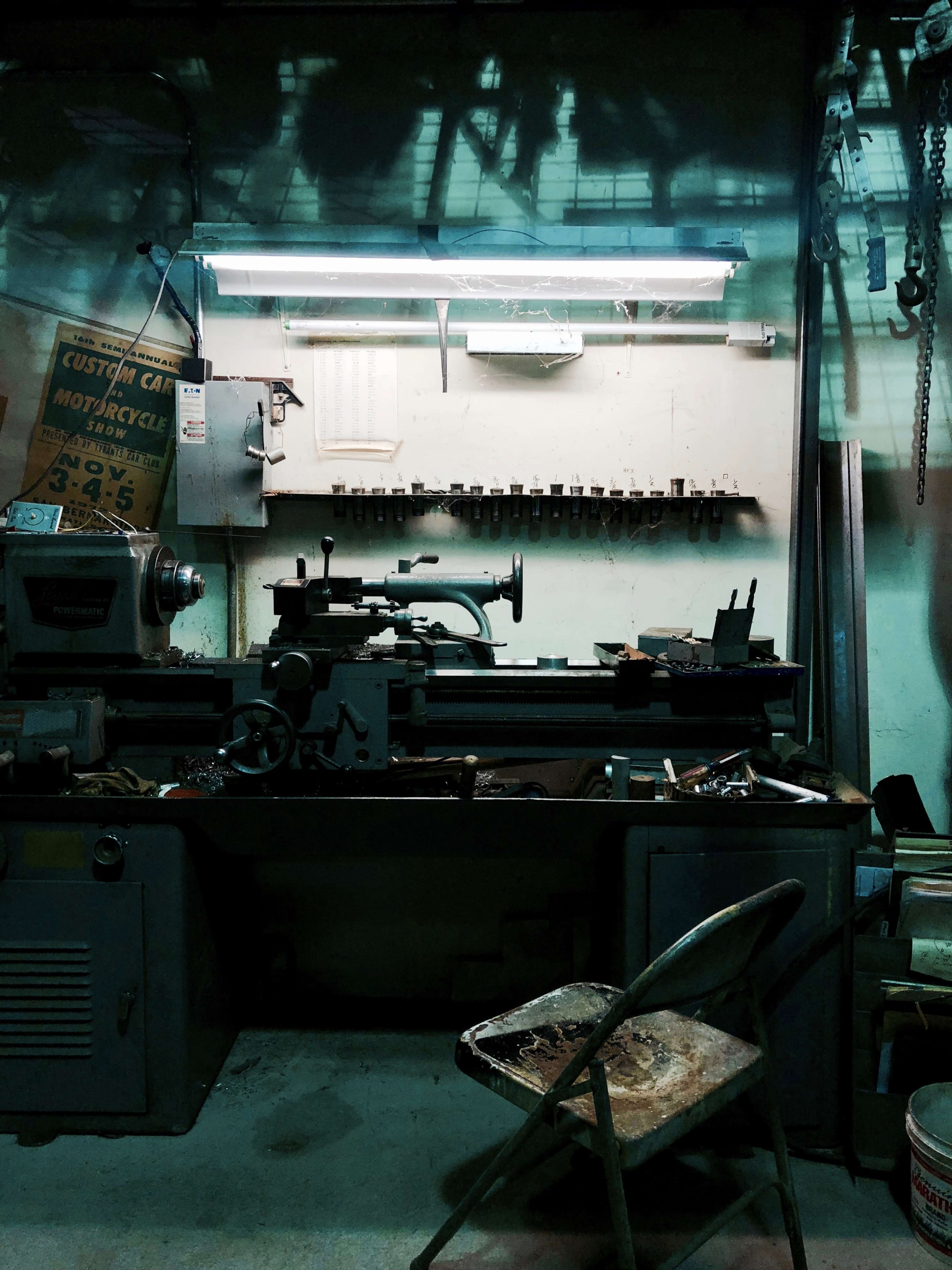 A dimly lit workshop with a lathe machine, tools on the wall, and a worn-out metal chair in the foreground.