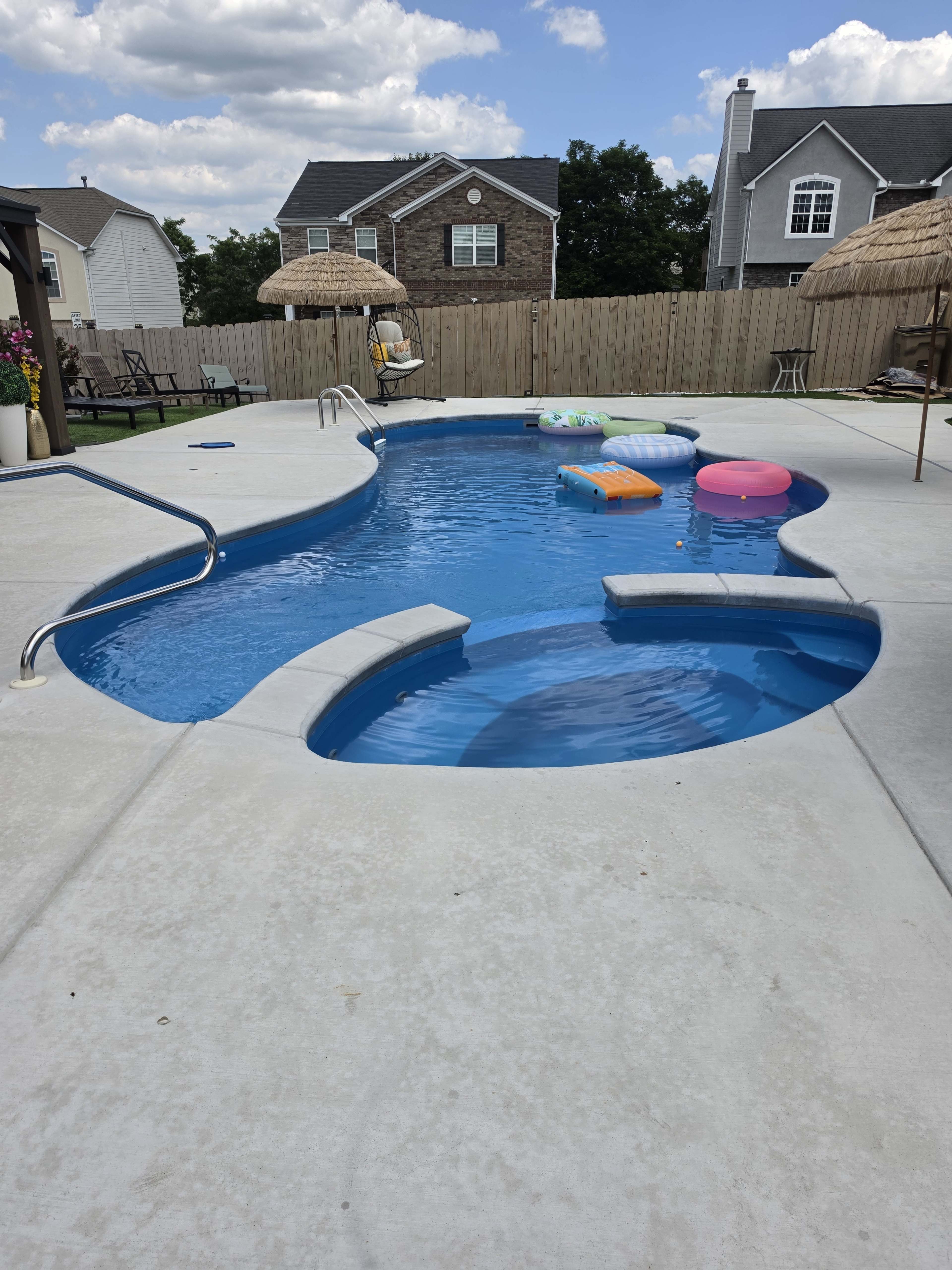 The image shows a rectangular swimming pool with colorful floaties, surrounded by a concrete deck and shaded by two straw umbrellas.