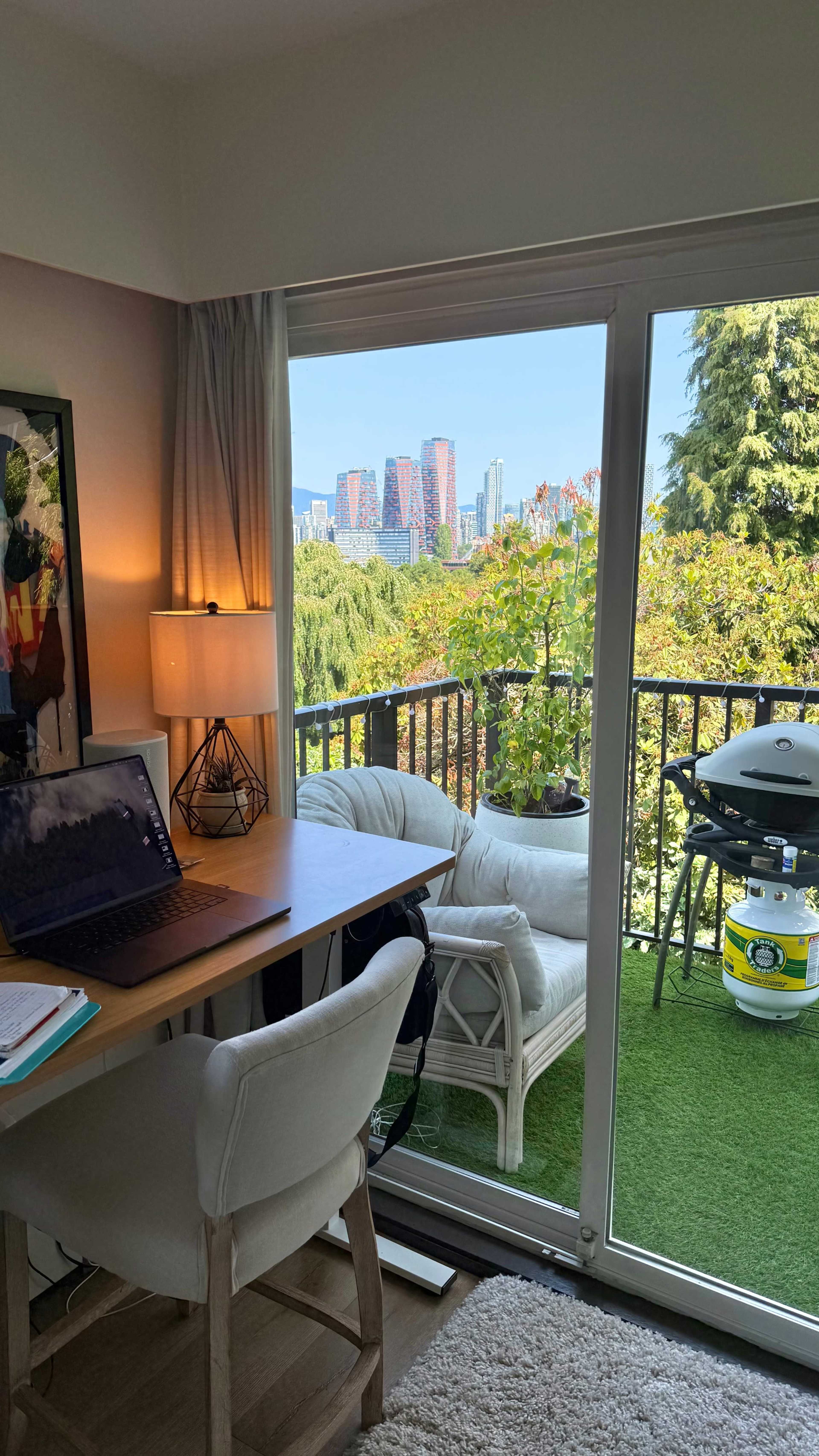 A workspace with a laptop and a chair overlooks a balcony with a grill and potted plants, framed by a view of high-rise buildings in the distance.