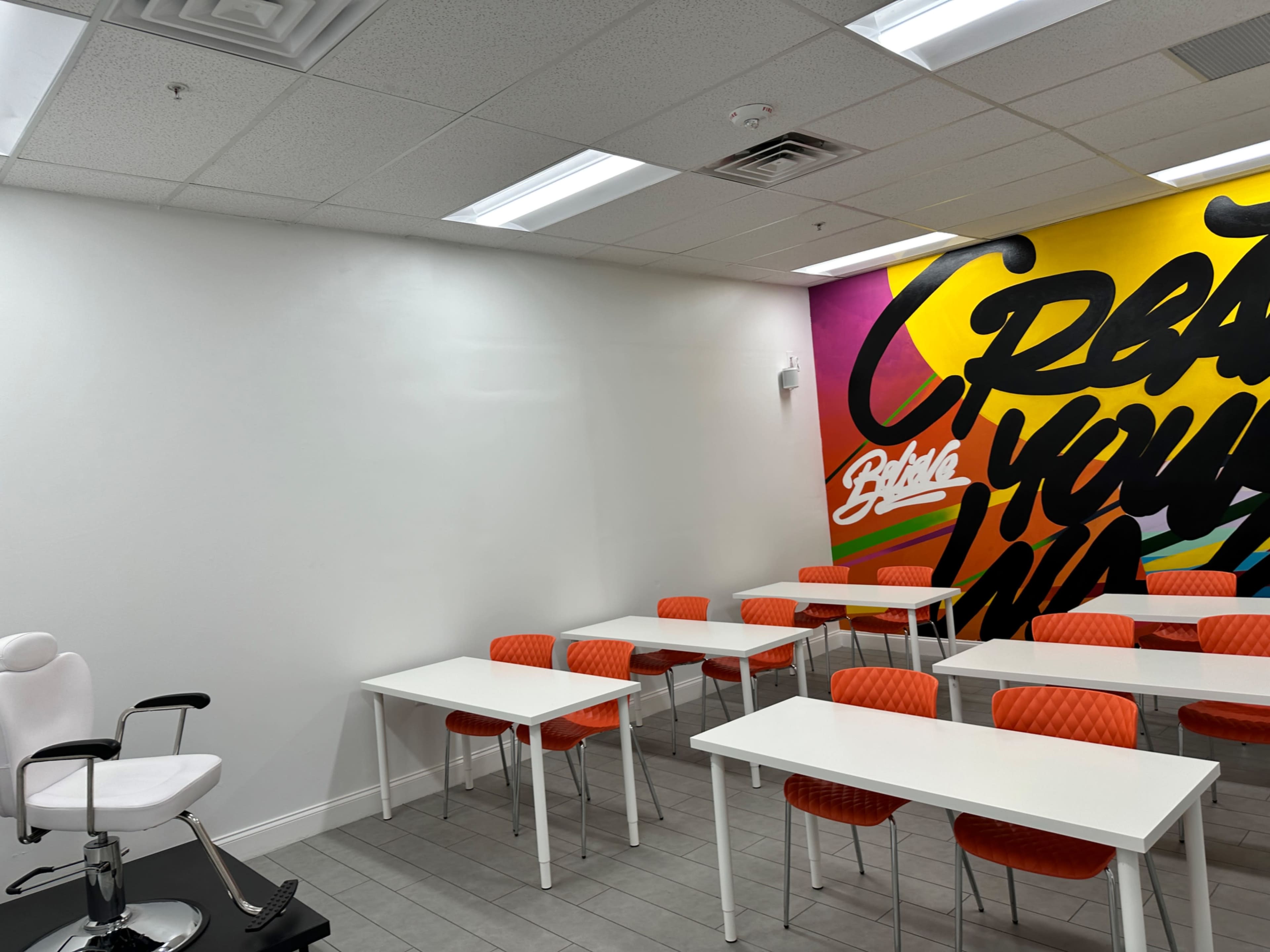 A bright classroom features orange chairs and white tables, with a colorful mural on one wall that reads "Create Your Own."