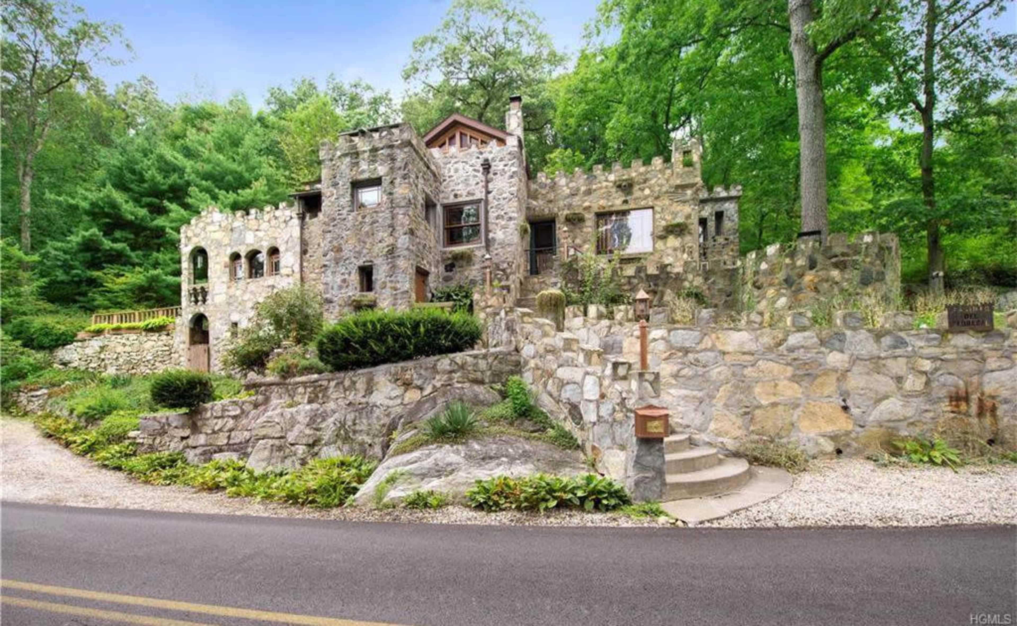 A stone castle-like house is set among trees, positioned on a hillside with a winding driveway leading up to it.