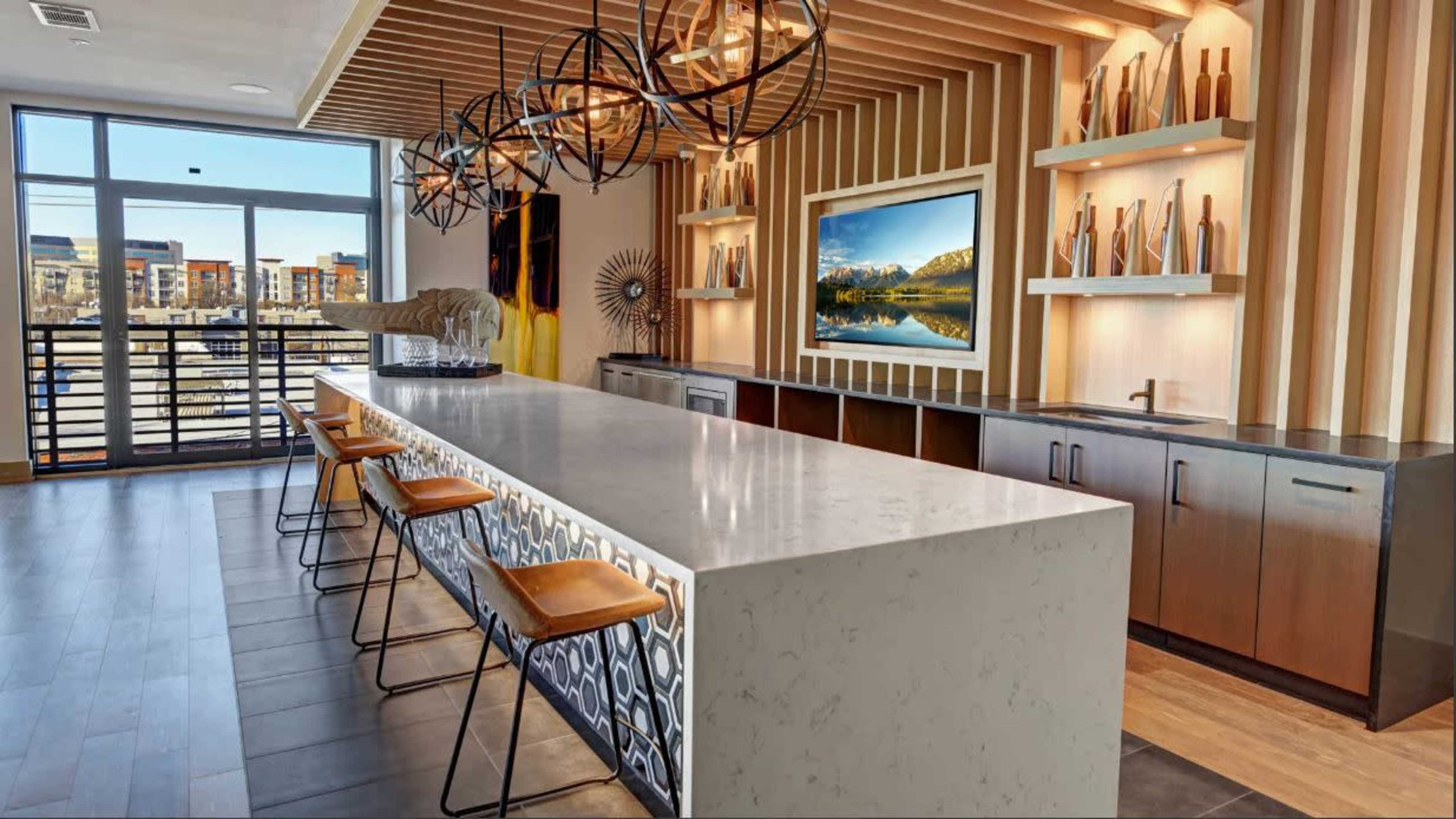 A modern kitchen with a long white countertop, beige bar stools, and decorative lighting fixtures above.