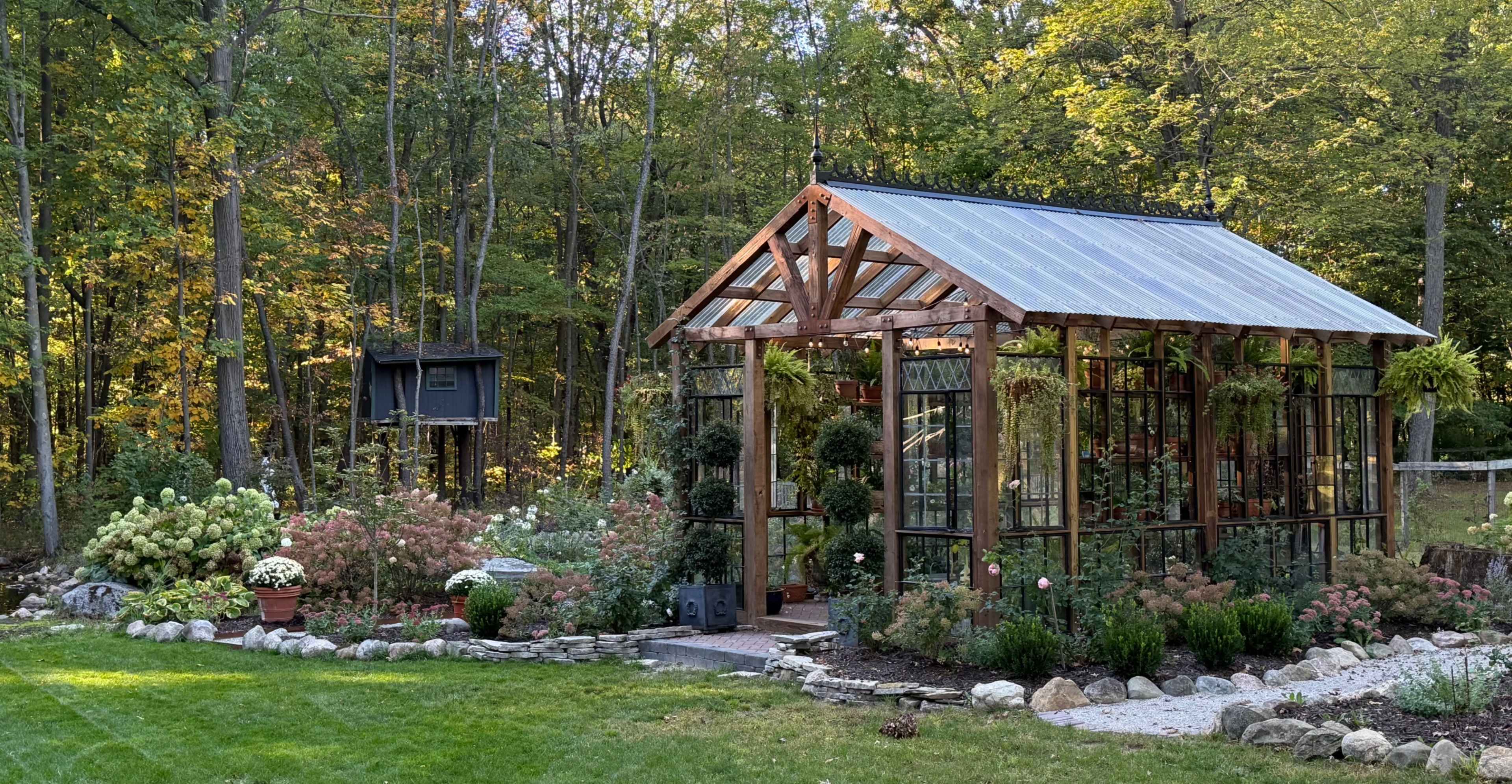 A wooden-framed greenhouse with glass panels is surrounded by a landscaped garden featuring various plants and flowers, set against a backdrop of trees.