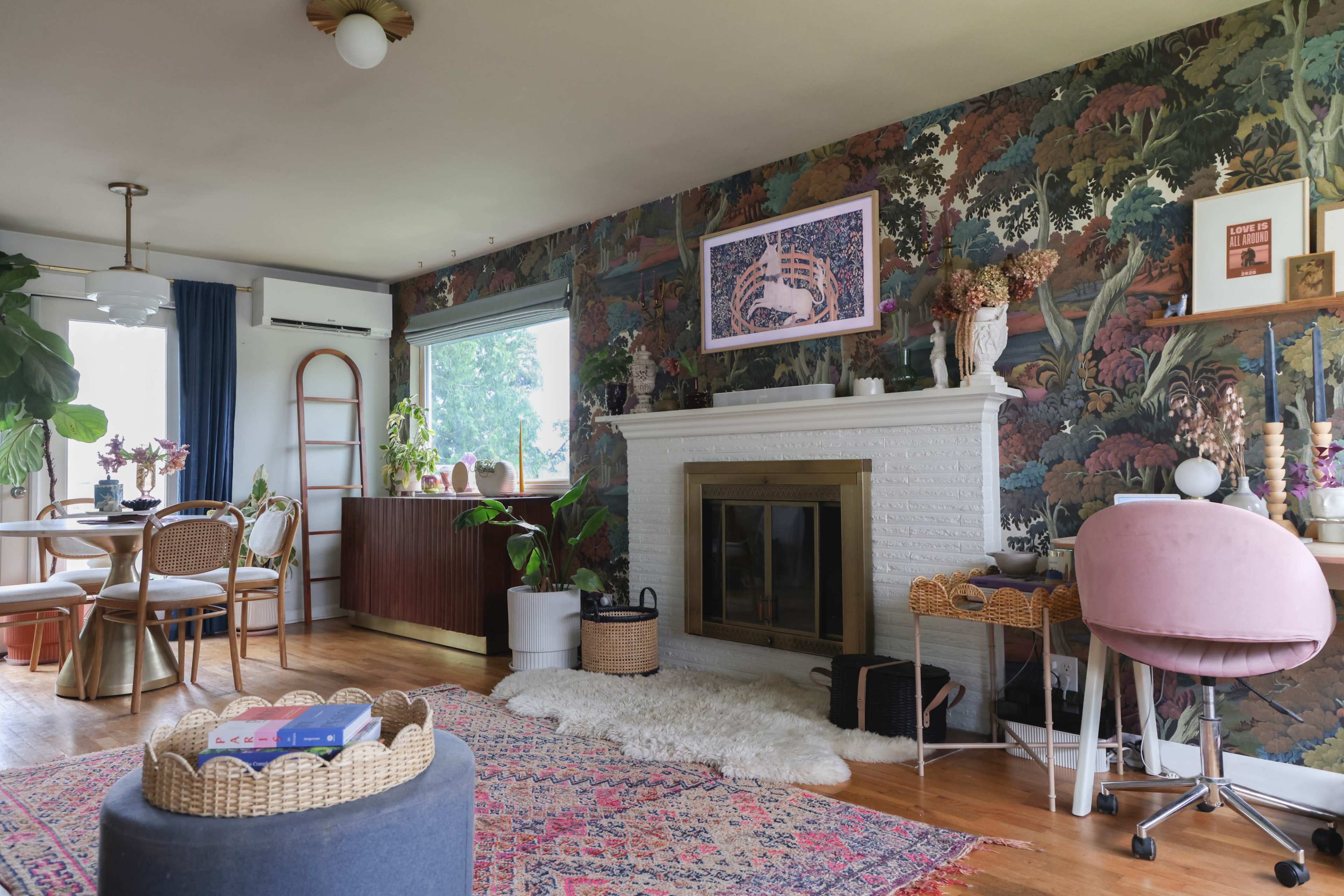 The living room features a decorative wallpaper, a white fireplace, and a combination of modern and vintage furniture, including a pink chair and a round ottoman on a textured rug.