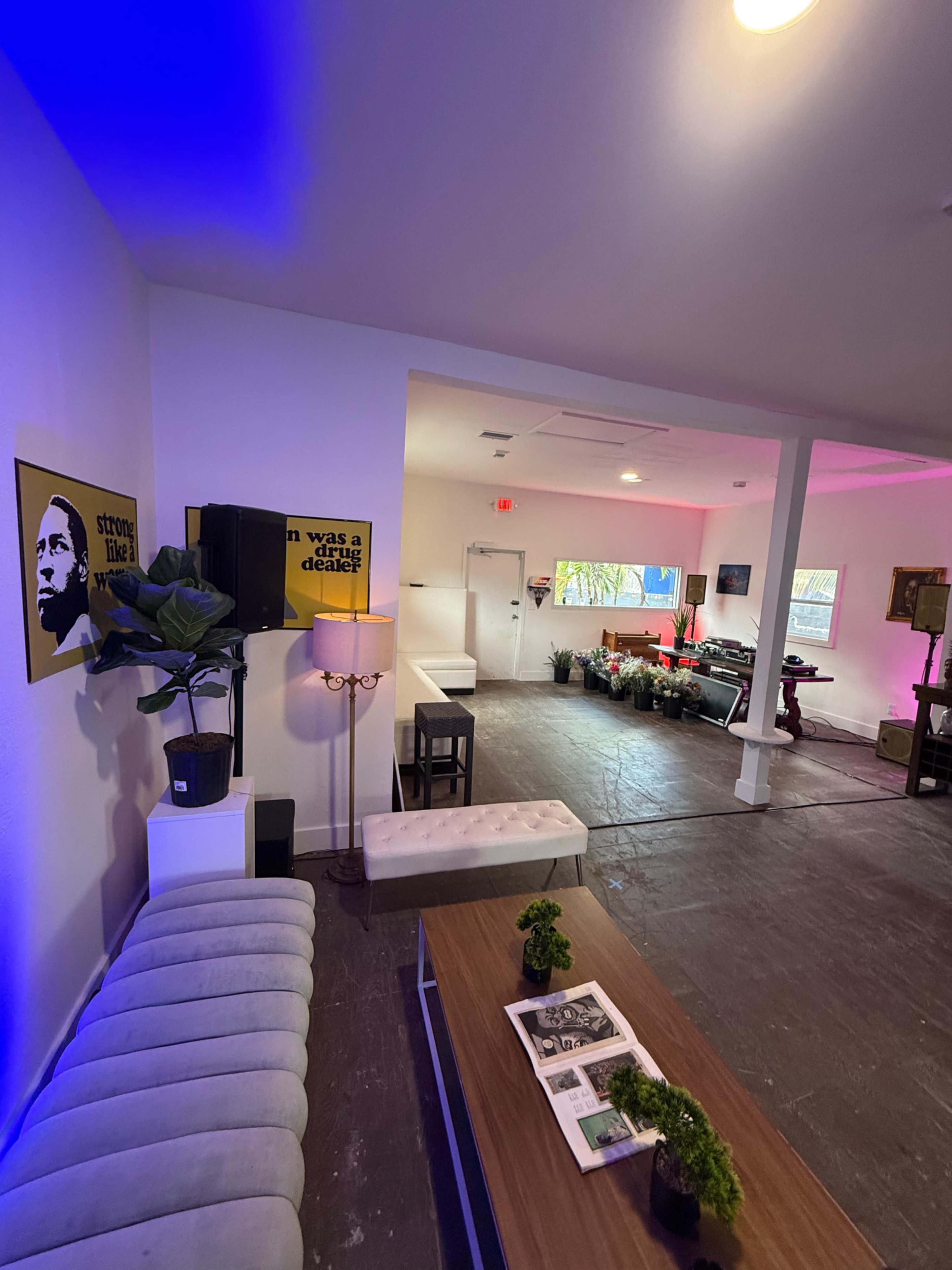 The image shows a modern, brightly lit interior space featuring a seating area with a couch, a coffee table, and a variety of plants, alongside artwork on the walls.