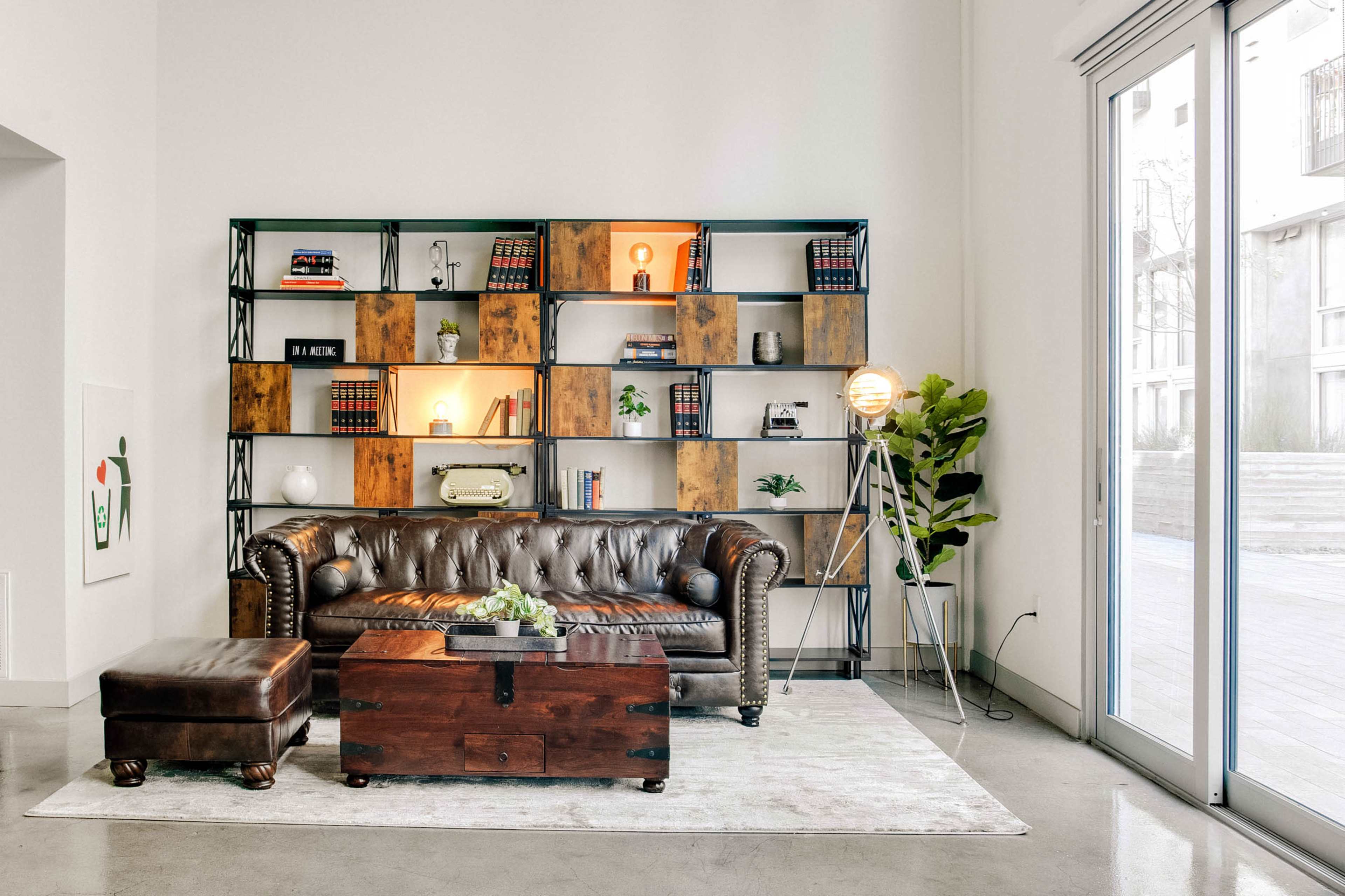 A leather sofa and a wooden trunk are arranged in a modern living room with a bookshelf and large glass doors leading to an outdoor area.