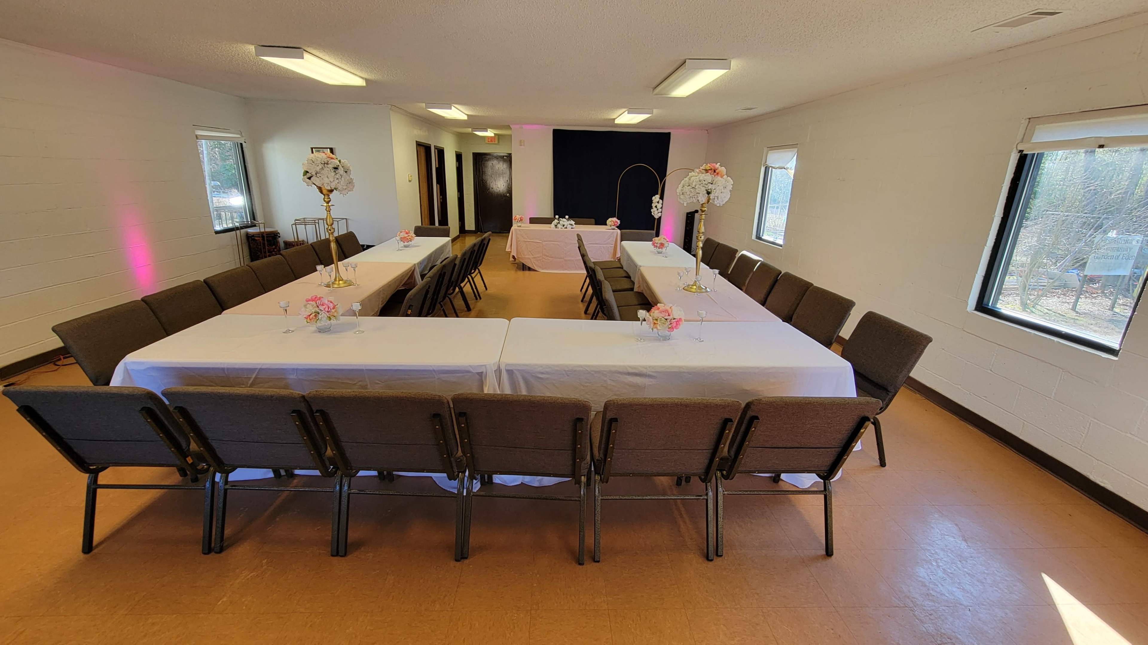 The image shows a spacious banquet hall set up with two long tables arranged in a U-shape, adorned with white tablecloths and small floral centerpieces.