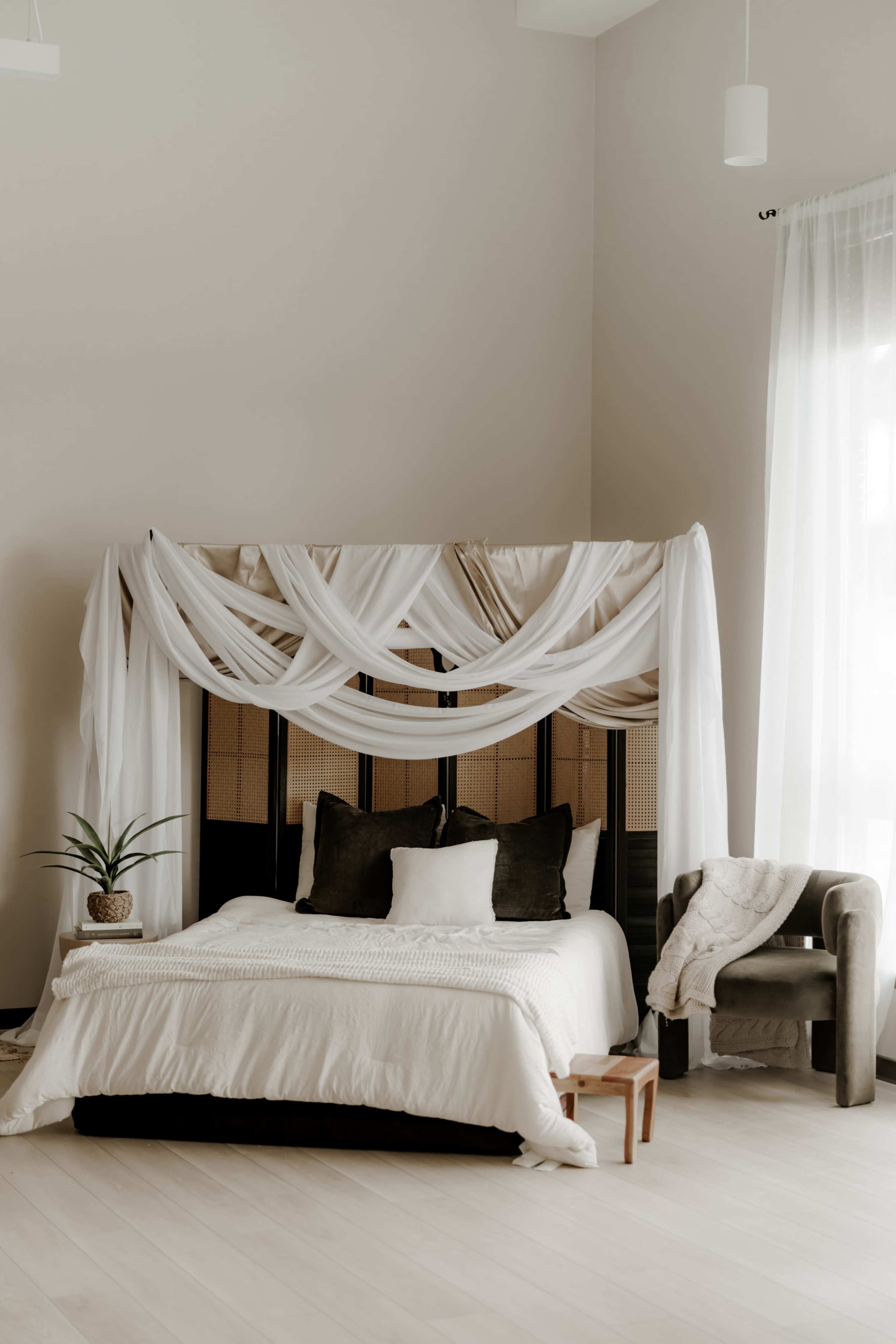 The image features a minimalist bedroom with a bed draped in white fabric, a potted plant, and a cozy armchair in a softly lit environment.