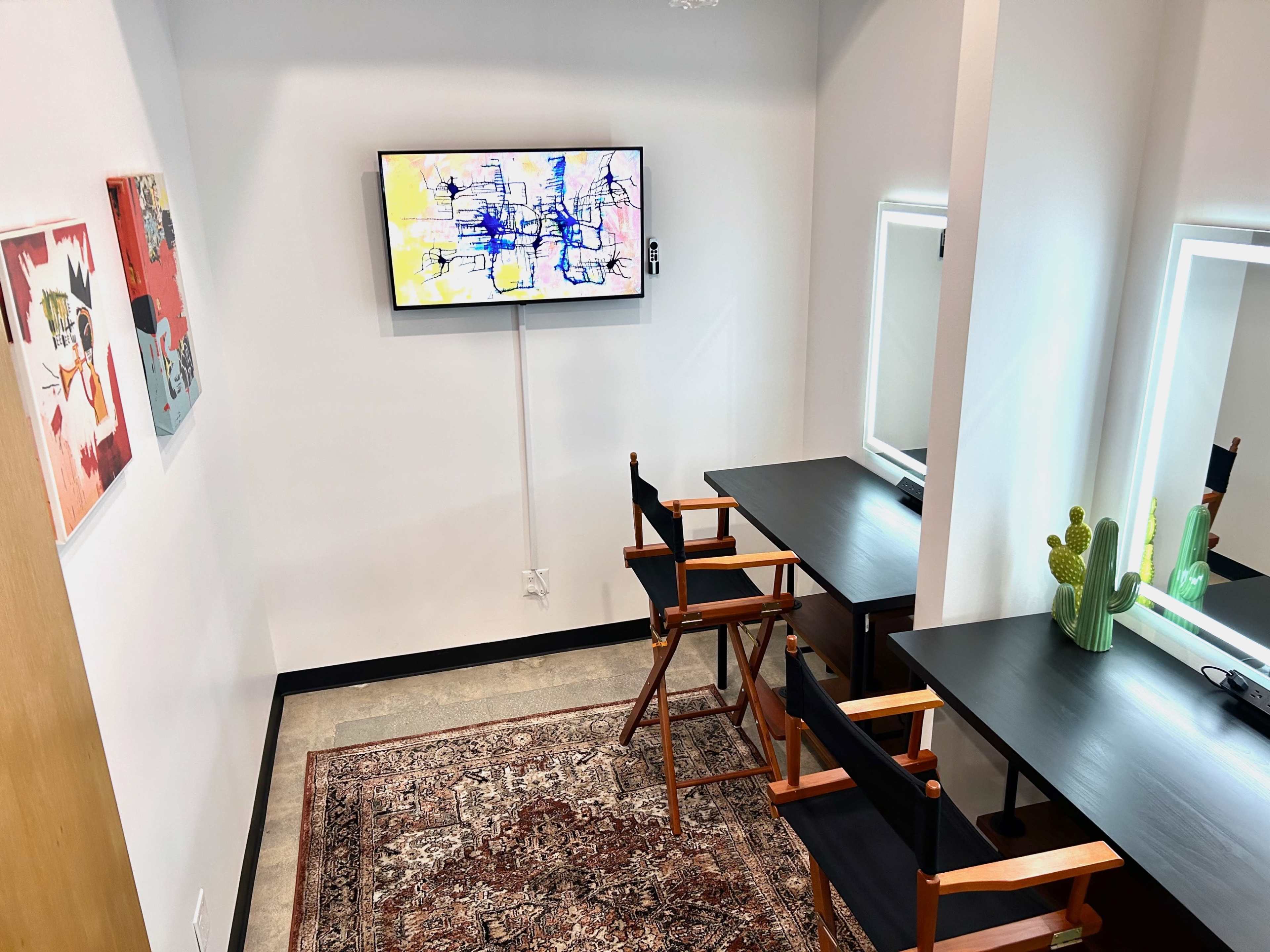 A small room with two director's chairs facing a wall-mounted television and mirrors, alongside colorful artwork and a rug on the floor.