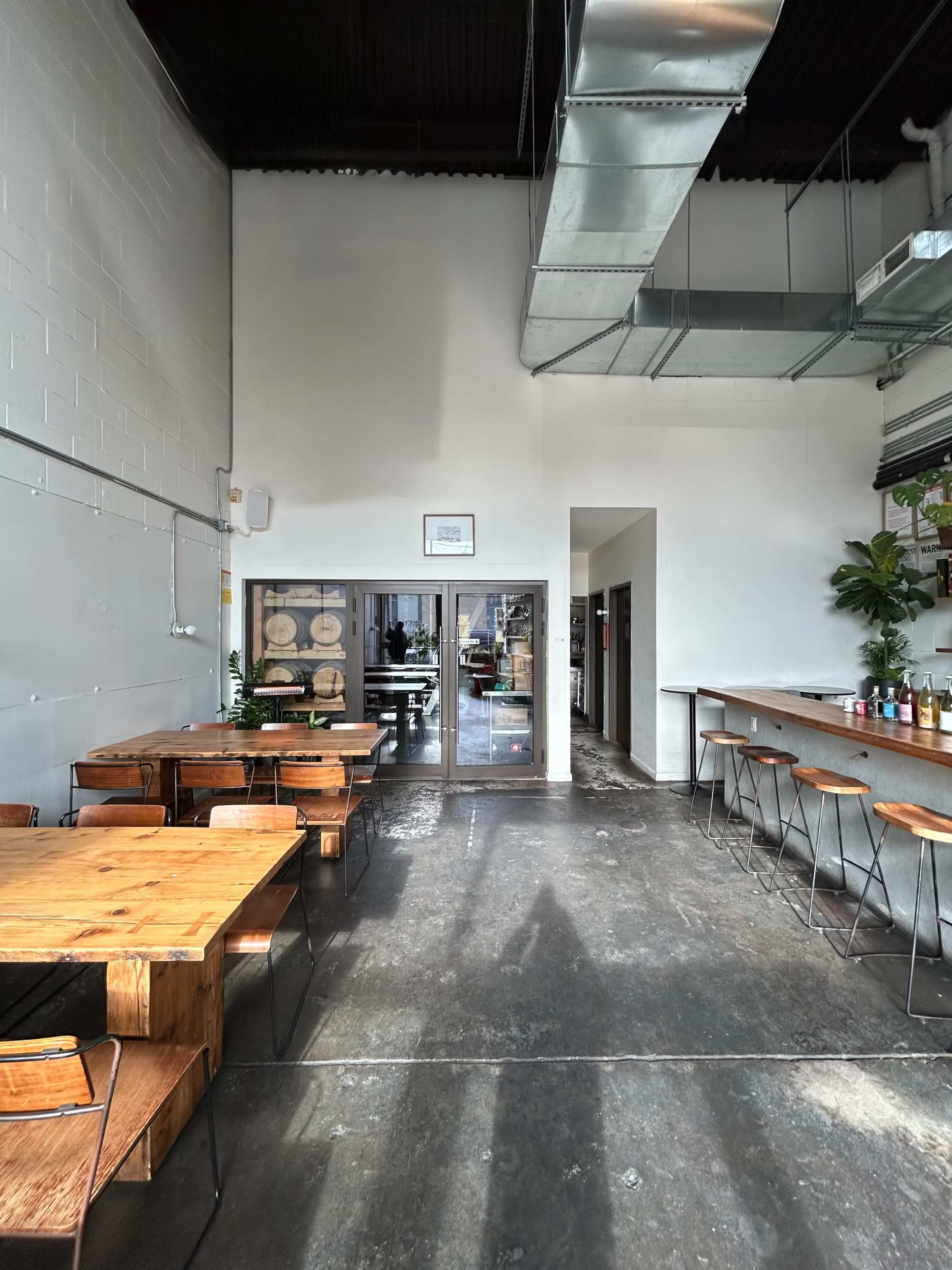 The image shows a modern, minimalist café interior with wooden tables, metal chairs, and a bar area, featuring large windows that overlook a workspace.