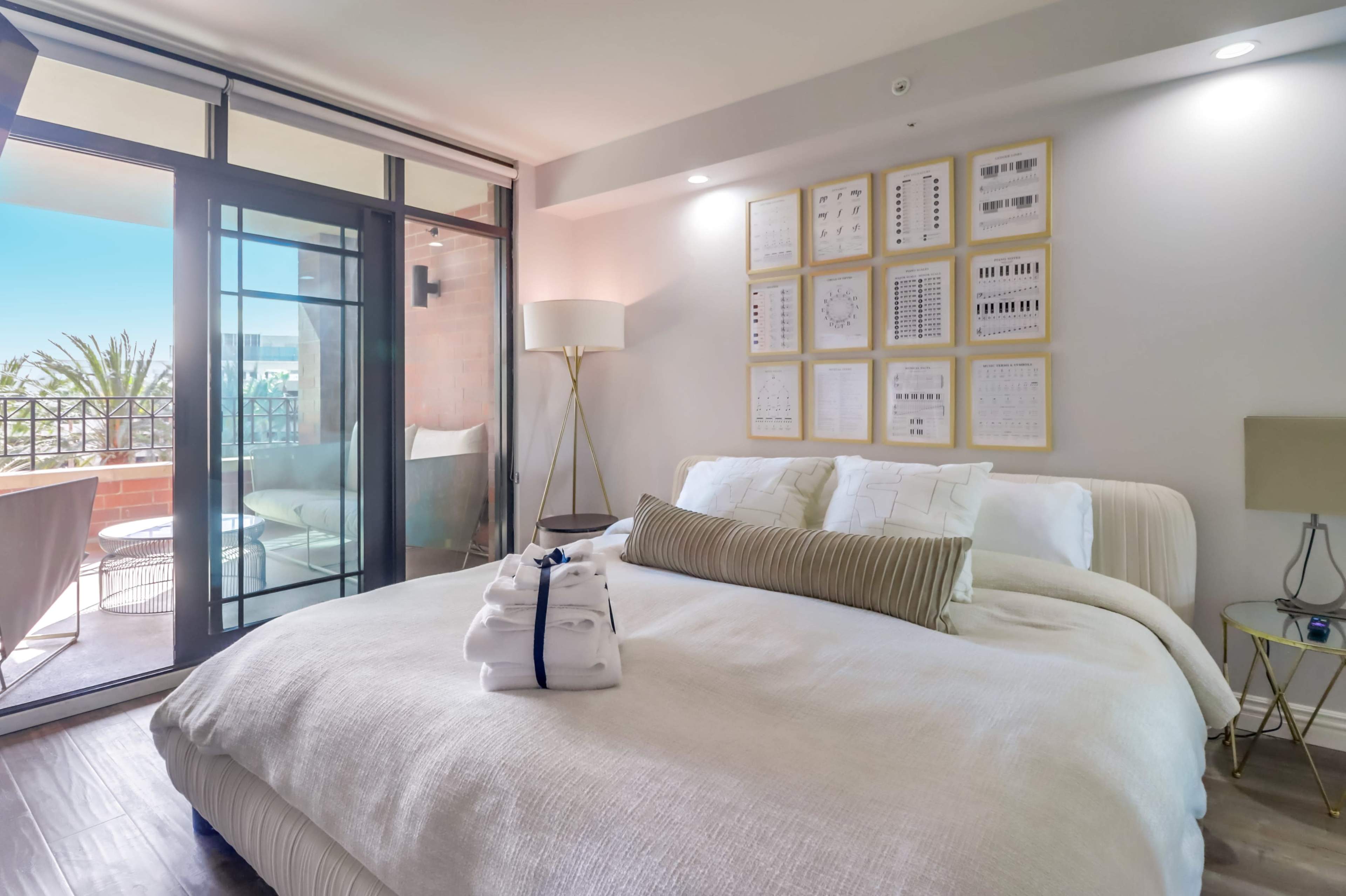 A modern bedroom with a large bed, decorative pillows, a lamp, and a balcony visible through a glass door.